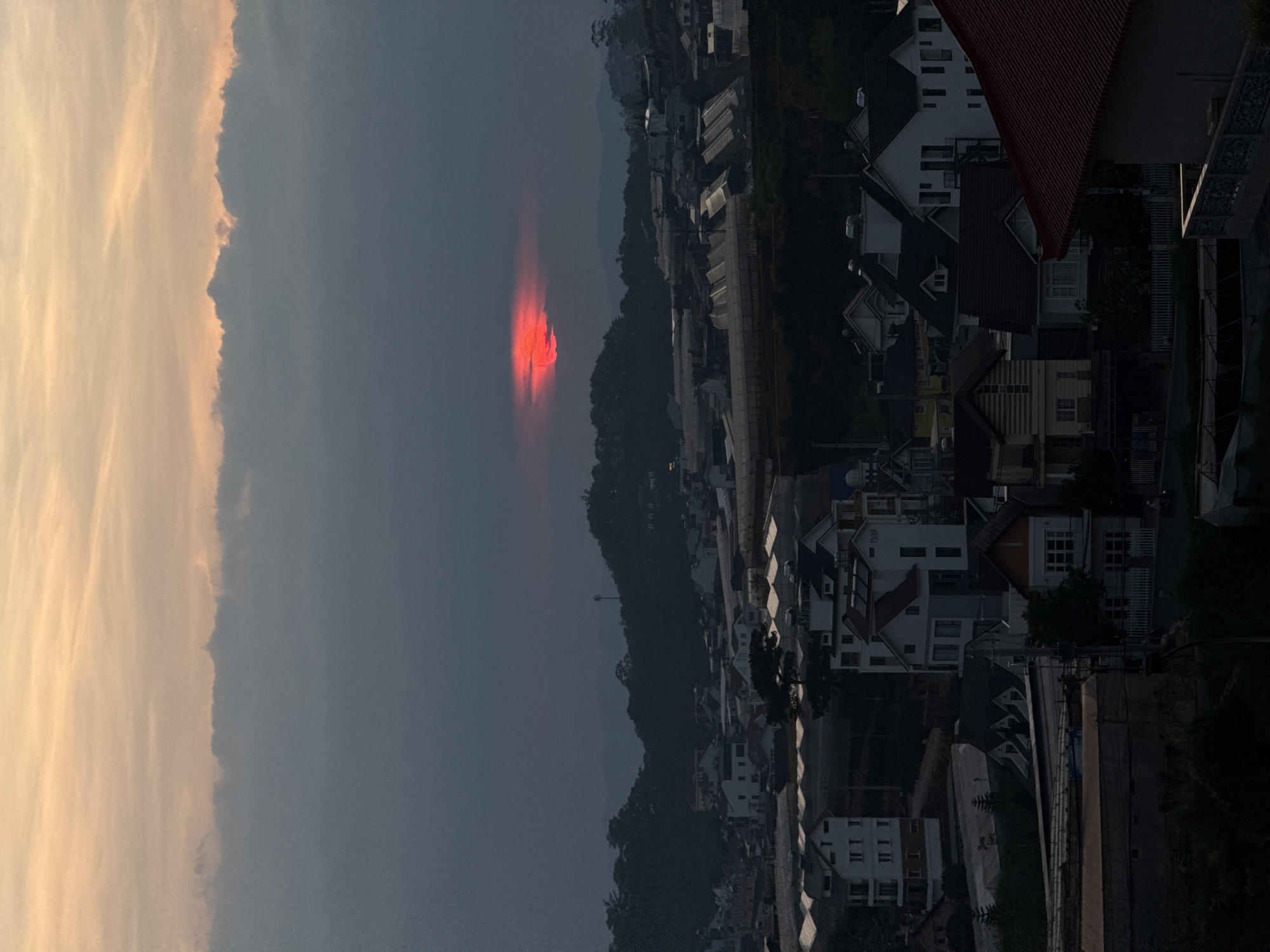 The image shows a landscape in Lâm Viên - Đà Lạt, Vietnam. In the distance, a bright orange sunset is visible, partially obscured by a hill. A layer of mist or fog hangs in the air. The foreground features residential buildings and structures, with diverse architectural styles and colors. Some buildings are brightly lit, while others are in shadow. A cloud formation with orange and yellow hues occupies the left side of the frame. The entire scene is captured in an overcast, dim setting.