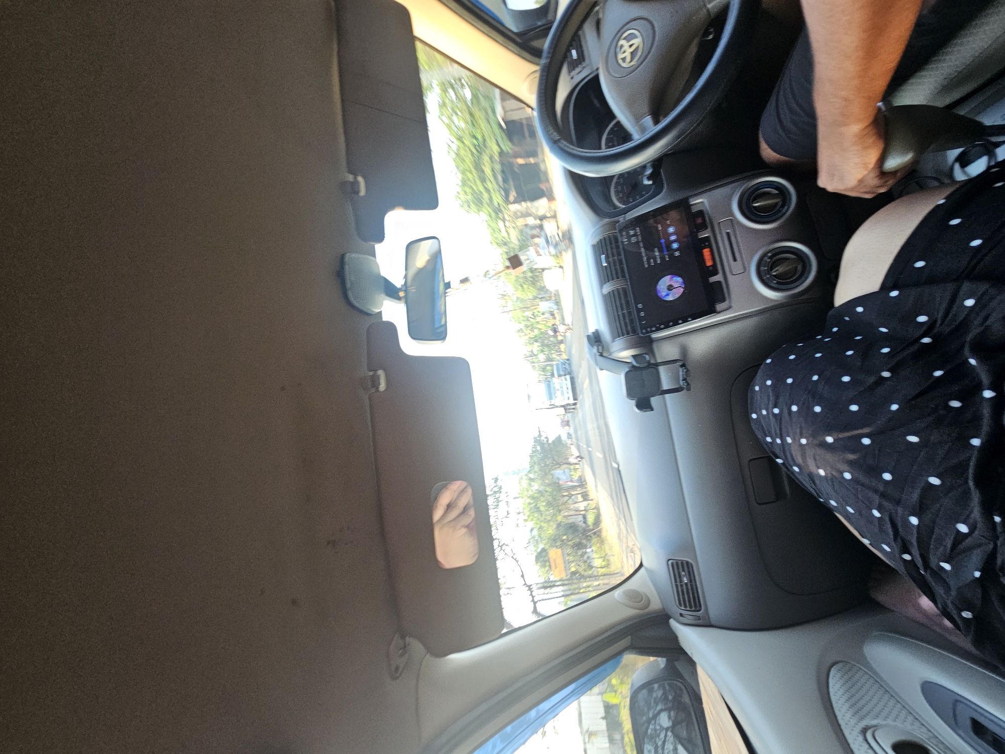 The image shows the interior of a car from the perspective of a passenger. The passenger in the foreground is wearing a black skirt with white polka dots. Their legs are visible. A person's arm and hand are on the gear shifter. The dashboard of the car is visible, including the steering wheel with a Toyota logo, the car's entertainment system displaying a colorful graphic, air vents, and climate control knobs. The view through the windshield shows a road with trees and buildings on either side, suggesting an urban or suburban environment. There are also vehicles visible on the road, including a truck in the distance. The sky is bright, indicating daytime. The image captures a moment during travel.