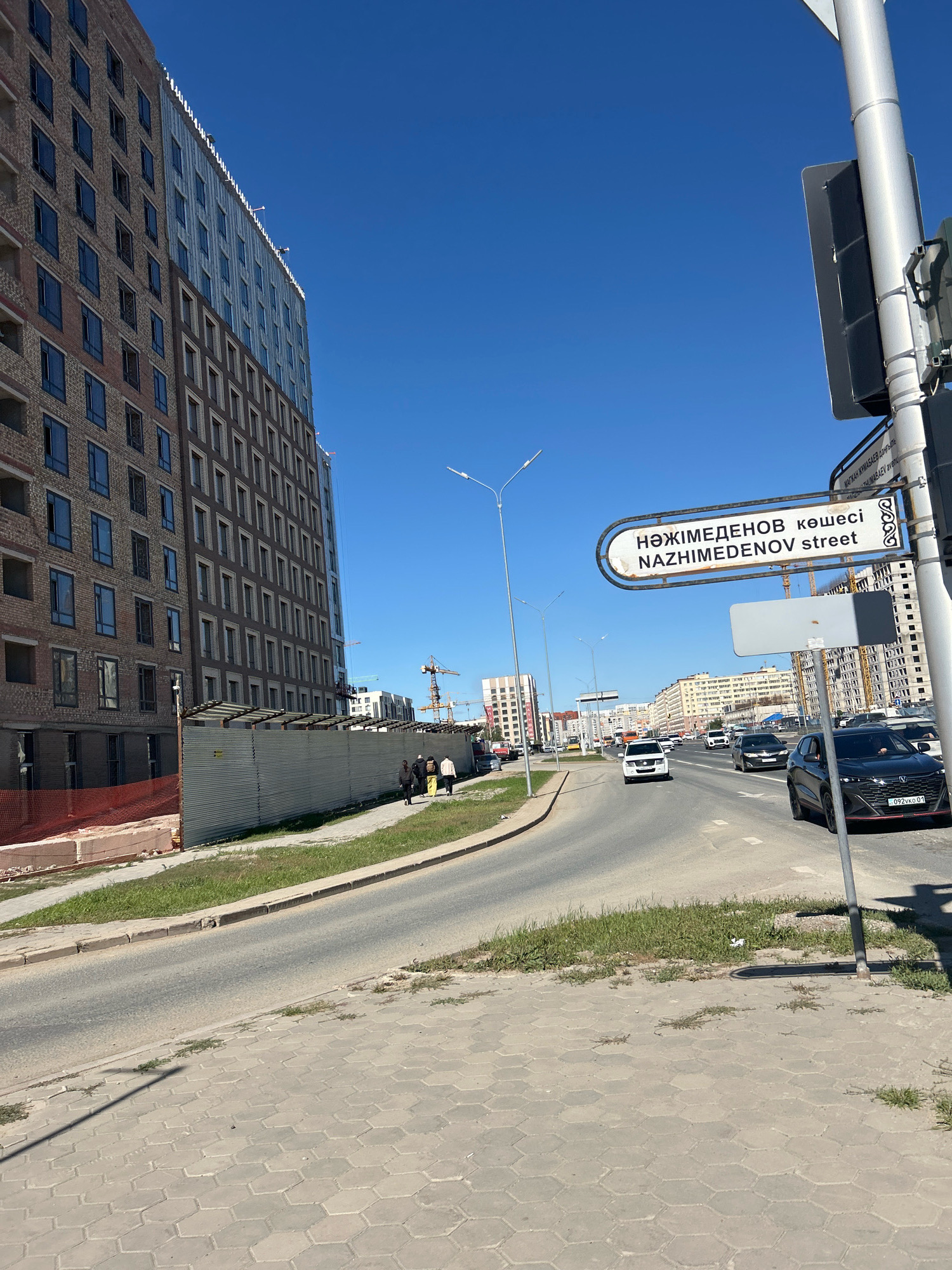 This is a photograph taken on a clear, sunny day in Astana, Kazakhstan. The image captures a street scene with several buildings under construction and completed. On the left side of the frame, a tall, multi-story building is visible, appearing to be in the midst of construction with exposed brickwork and unfinished windows. A metal fence with some red netting is also present in front of this building.
Further down the street, a signpost displays street names in both Kazakh and English: "НӘЖІМЕДЕНОВ көшесі" and "NAZHIMEDENOV street". Several cars are driving on the road, including a black SUV in the foreground with a license plate reading "092vko 01". In the middle ground, more buildings and traffic are visible. The sky is a bright, clear blue. Three people are walking on the sidewalk in the middle ground, moving away from the camera. A traffic light is partially visible on the right side of the frame. The ground is paved with hexagonal concrete tiles in the foreground.