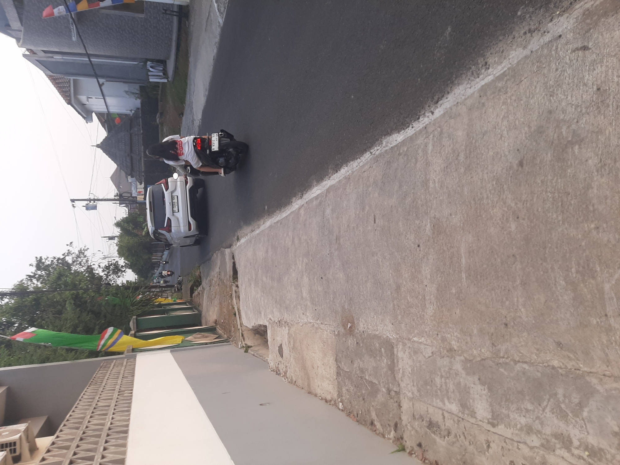 The image captures a street scene in Sumampir, Indonesia, with a focus on the side of the road and a passing motorcycle. On the left, a textured white wall borders a concrete sidewalk that appears weathered and uneven, with a small, broken drainage channel visible. Beyond the sidewalk, a green fence runs along a property. In the background, trees and residential buildings line the street. A white car is parked further down the road, and a motorcycle with two occupants is in motion, traveling away from the viewer. The rider, wearing a white t-shirt with a graphic, is in front, and a passenger sits behind. The motorcycle has a license plate that reads "N 6312 UJ". The overall lighting suggests it is either early morning or late afternoon.