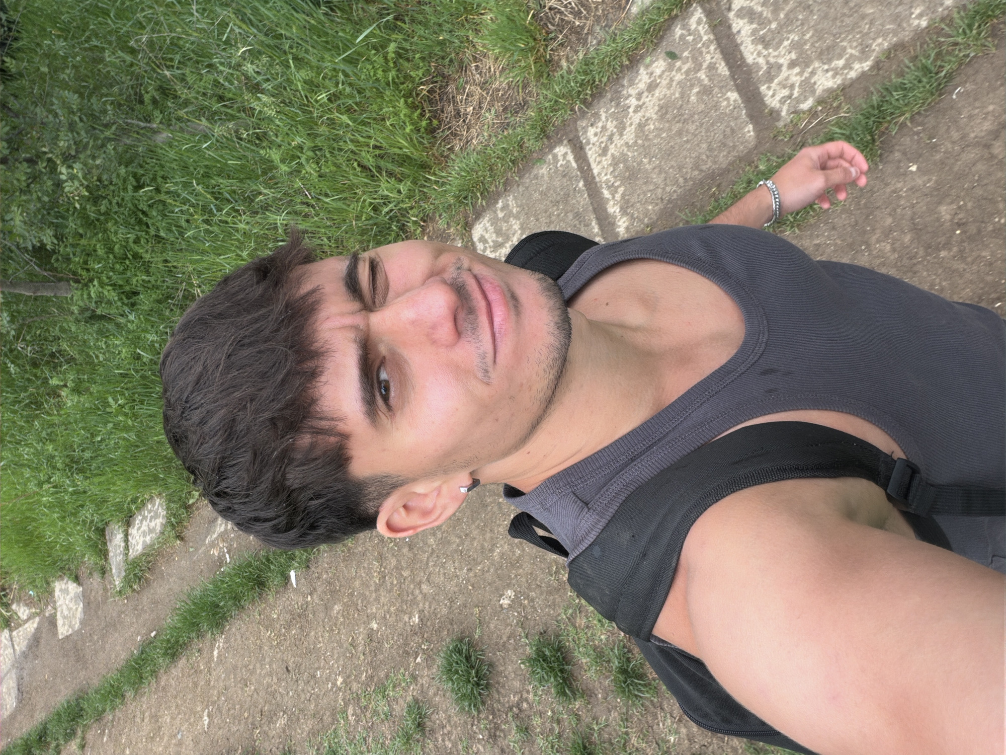A selfie shows a man with dark, wavy hair and a trimmed beard. He is wearing a dark gray tank top and a black backpack. He has a silver earring in his left ear and a silver bracelet on his left wrist. He is standing on a dirt path next to a concrete walkway. Grass and some shrubbery is nearby. The location is not discernible as Sofia, Bulgaria, based on the provided details.