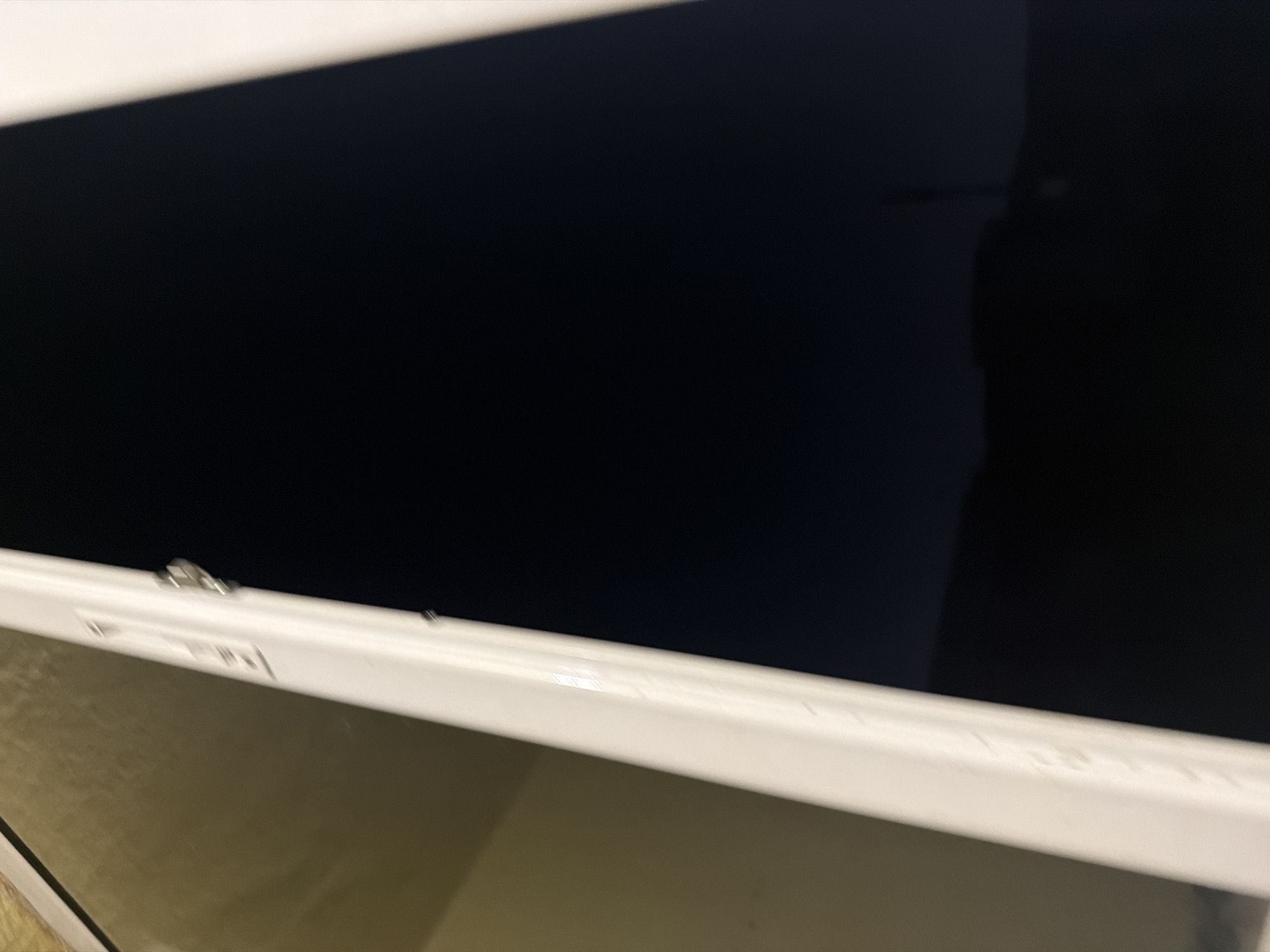 The image presents a forensic view through a window or glass door frame at night in Sofia, Bulgaria. A prominent white frame, likely part of a UPVC window, traverses the image diagonally from the upper left to the lower right, separating an interior reflection from an exterior view. On the left, the glass surface reflects a blurred, yellowish-beige interior, indicative of an illuminated indoor space, possibly a wall or ceiling. On the right, the glass reveals a very dark exterior, characteristic of nighttime. Within this dark reflection, the faint, indistinct silhouettes of distant hills or low-lying terrain are discernible towards the bottom. Further up in the dark expanse, a slender, vertical structure, potentially a distant antenna or tower, is barely visible. A small, faint, circular light source also appears in the lower-middle section of the dark area, suggesting a distant artificial light. The white frame itself exhibits minor surface imperfections and houses a metallic locking or hinge mechanism near its top. No people or specific actions are visible; the scene focuses on the architectural elements and the ambient nocturnal environment.