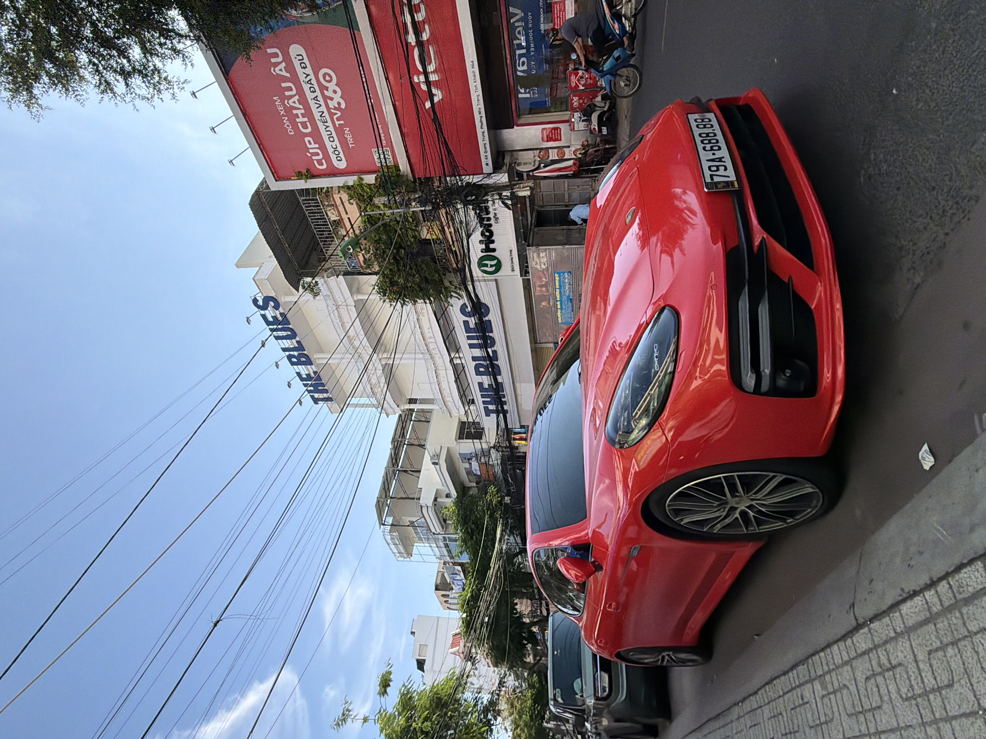 A low-angle, eye-level, full shot captures a red Porsche Panamera parked on the side of a city street in Nha Trang, Vietnam. The car's front is positioned facing the right, with its hood and headlights visible. Its license plate reads "79A-688.88". Next to it, a dark gray SUV is also parked. The street is paved, and a sidewalk with patterned tiles is to the left of the vehicles.
Behind the cars, a multi-story building with a white facade is visible, featuring balconies and rooftops. Signage on the building includes "THE BLUES" in large blue letters. To the right of "THE BLUES," a large red billboard displays text in Vietnamese, with a prominent "TV360" logo. Another building to the right has "VIET" in large red letters.
The sky above is clear and blue with some scattered white clouds and a network of overhead electrical wires crisscrossing the scene. Trees with green foliage line the street and buildings, adding to the urban landscape. In the background, a person rides a motorcycle on the street. The lighting suggests it is daytime, likely late afternoon due to the soft shadows.