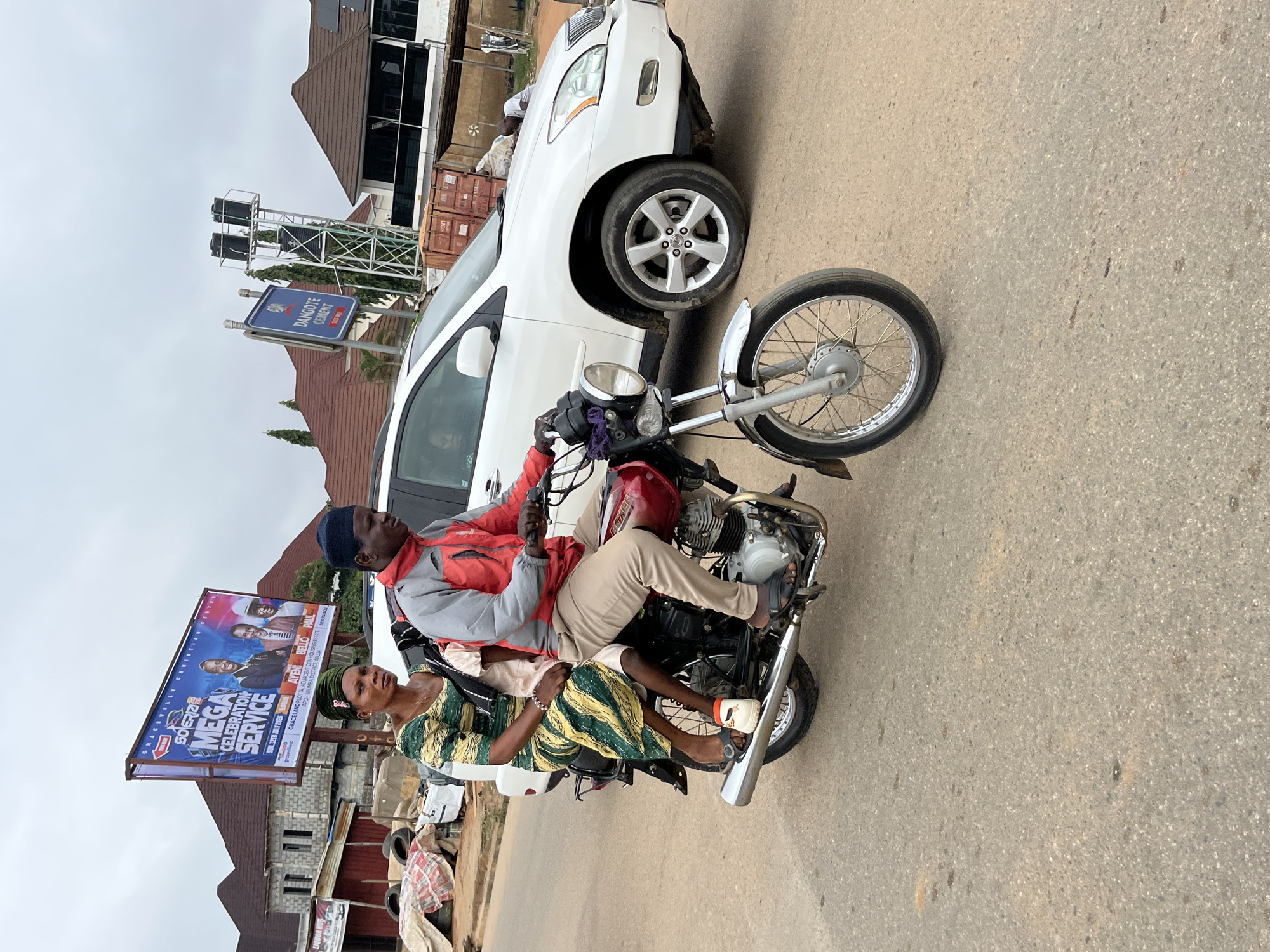 An overcast day in Abuja, Nigeria, showcases a three-person motorcycle caravan. The motorcycle, a red Boxer, is ridden by a man in a red and gray jacket and khaki pants, with a black beanie. A woman in a green and yellow patterned dress sits behind him, and a child, also in a pink dress, is seated between them. The trio is captured mid-journey on a paved road. In the background, a white SUV is parked to the right, with a "Dangote Cement" billboard and a tall structure with water tanks visible further beyond. The scene is set with various buildings and some discarded materials on the side of the road, suggesting a bustling urban environment.