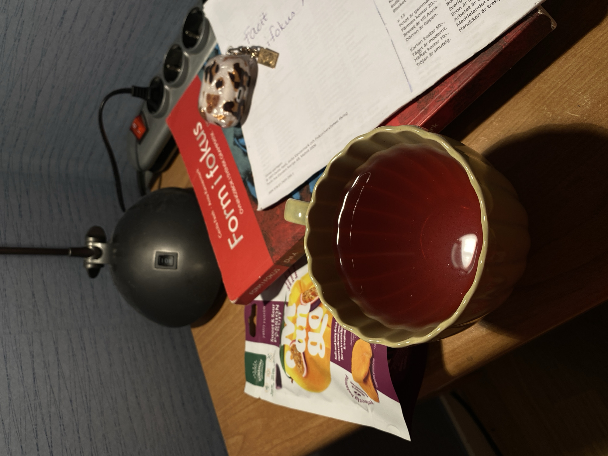 The image shows a desk or table with several objects. A cup of red liquid (possibly tea or juice) sits on the right side of the frame, partially obscuring a packet of "Fruta Cub" sweet and sour passion fruit jelly candies. Adjacent to the candies is a red book titled "Form i Fokus" (Form in Focus), which appears to be a language learning workbook. A piece of white paper with handwritten notes ("facit fokus") rests on top of the book. A small, decorative object (possibly a charm or keychain) is on the paper. To the left is a black desk lamp and a power strip. The background features a blue patterned wall. No specific people or location in Björklinge, Sweden, can be identified from the image.