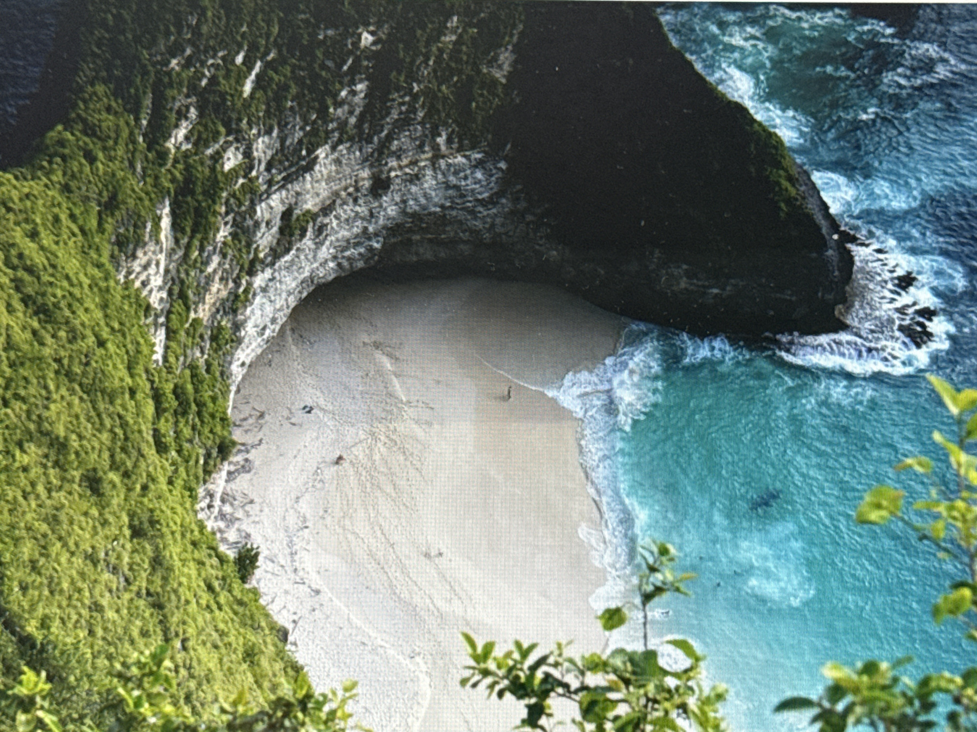 This is an aerial, high-angle shot of a secluded beach nestled within a cove. Lush green vegetation covers steep, rocky cliffs that curve around the sandy shore. The ocean water is a vibrant turquoise, with white waves crashing against the rocks and receding onto the beach. The sand itself appears light-colored and marked with lines, possibly from receding tides or footprints. A solitary figure can be seen walking on the beach, and a few more individuals are visible in the water. The overall scene depicts a beautiful, natural coastal landscape. This image is not located in Pushkino, Russia; it depicts a tropical or sub-tropical coastal environment.