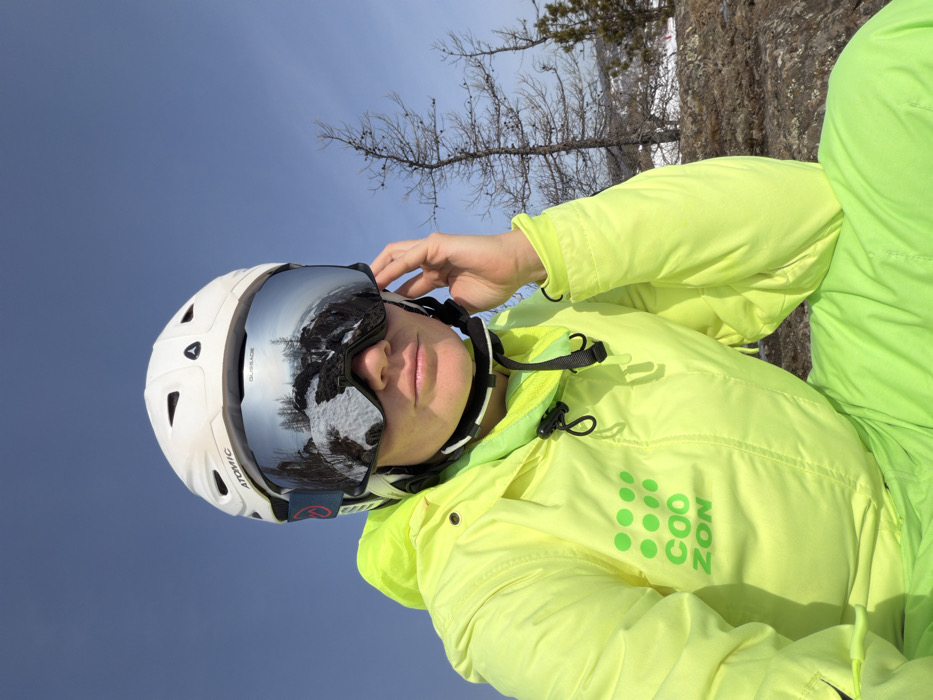 An adult female individual is depicted in an outdoor, elevated winter environment in Zelenaya Polyana, Russia. She is positioned facing the camera, wearing a white "ATOMIC" brand ski helmet and large, mirrored "GLASSLADE" brand ski goggles. The goggles reflect a snowy, rocky, forested landscape under a clear blue sky. She is dressed in a neon yellow/lime green ski jacket with a hood, featuring a dark green "COO ZON" logo and dot pattern on the left chest, and matching ski pants. Her left hand is raised to the right side of her head, with fingers near the temple area, seemingly adjusting the goggle strap. The background consists of sparse, bare trees and rocky ground under a clear blue sky.