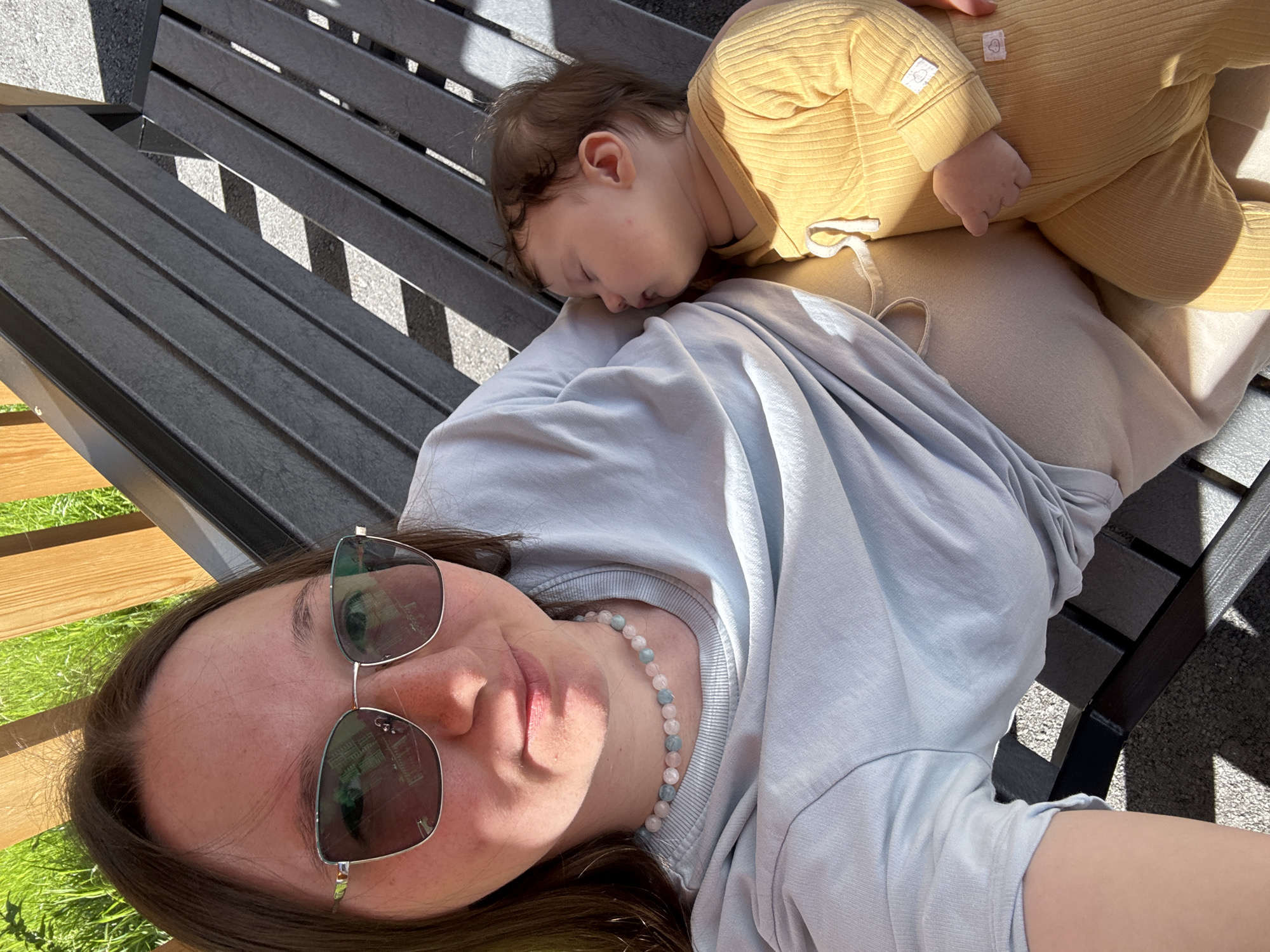 In this high-angle, close-up selfie, a woman and a baby are lying on a dark-slatted park bench. The woman is in the foreground, wearing sunglasses and a light blue long-sleeved shirt. A beaded necklace with pink and blue stones rests on her neck. Her expression is relaxed, with her eyes closed and lips slightly parted, suggesting a peaceful moment. The baby, dressed in a yellow ribbed onesie, is asleep on the woman's chest, nestled against her. The baby's face is partially obscured, but its peaceful slumber is evident. The scene is bathed in bright sunlight, casting sharp shadows from the bench slats onto the ground, indicating it's likely daytime on a clear day. Green grass and what appears to be a wooden bench are visible to the left of the main subjects. The overall mood is serene and intimate, capturing a quiet moment between the woman and the baby outdoors. The location context provided is Patrusheva, Russia.
