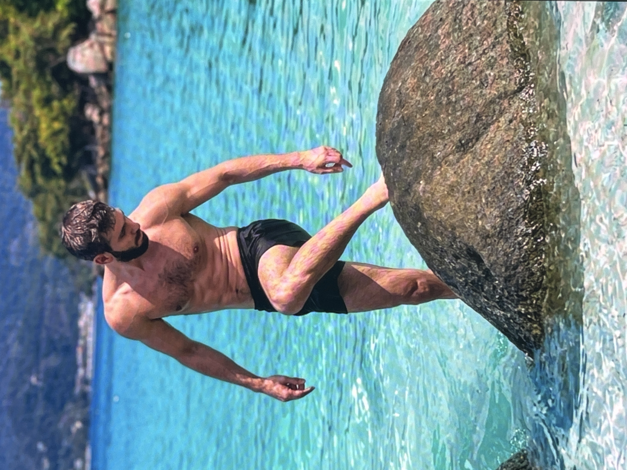 A shirtless man with dark hair and a beard stands in the turquoise water of a tropical ocean, facing to the right. He is wearing black swim trunks and has his right leg bent at the knee, with his foot resting on a large, dark, rounded rock in the foreground. His left arm is bent at the elbow and extended downwards, with his hand open, and his right arm is bent at the elbow and extended forward, with his fingers pointing downwards. The water is clear and shows the sandy bottom in some areas. In the background, there is a rocky, tree-covered coastline. The sky is not visible.