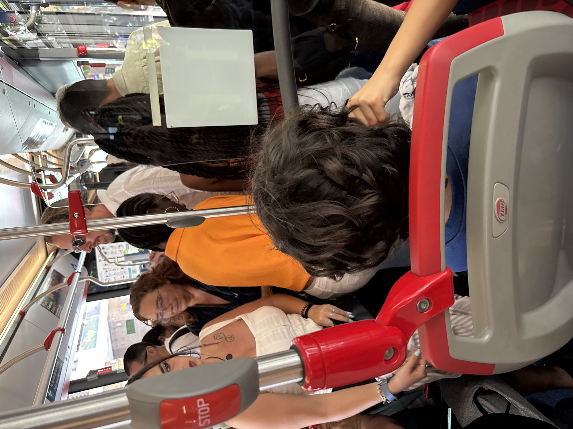The image depicts a crowded interior of a bus in Barcelona, Spain. Several passengers are standing and holding onto rails for support. One woman is wearing a white top and glasses, and another woman is wearing an orange shirt. A person wearing a blue shirt is partially visible near a red and gray bus seat. Braided hair is visible on another passenger. A "STOP" button on a handrail is also visible. The reflections in the bus windows show buildings outside, giving the impression that the bus is in motion.