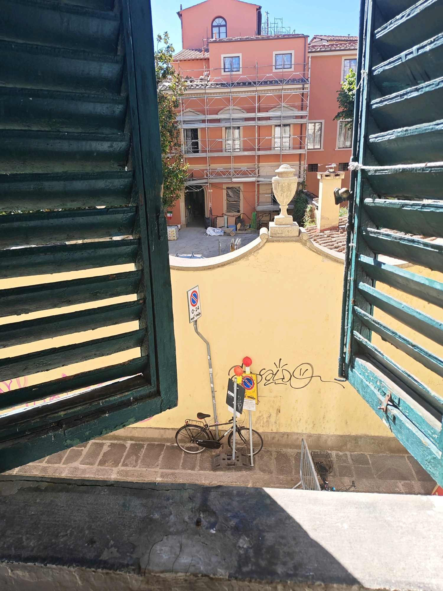 The image depicts a view through open, dark green wooden shutters, looking out onto a narrow street and buildings in Florence, Italy. The shutters themselves are weathered and show signs of peeling paint. The scene outside is bathed in bright sunlight.
In the mid-ground, a yellow stucco wall is visible. Graffiti in black ink, including a smiley face and stylized lettering, adorns the wall. To the left of the wall, a black bicycle is parked. Attached to a pole in front of the wall are several traffic signs, including a parking sign with the text "0-24". There are also what appear to be temporary traffic lights, one with red and yellow lights. A metal barrier is positioned in front of the bicycle.
Beyond the yellow wall, parts of buildings are visible. One building is partially covered in scaffolding, suggesting construction or renovation work. This building is painted in a peachy-orange hue and has several windows. Another building to its right is a similar color. Above these buildings, a clear blue sky is visible. The ground in front of the yellow wall is paved with stone or brick. In the foreground, the stone windowsill of the building from which the photo is taken is visible, along with shadows cast by the shutters.