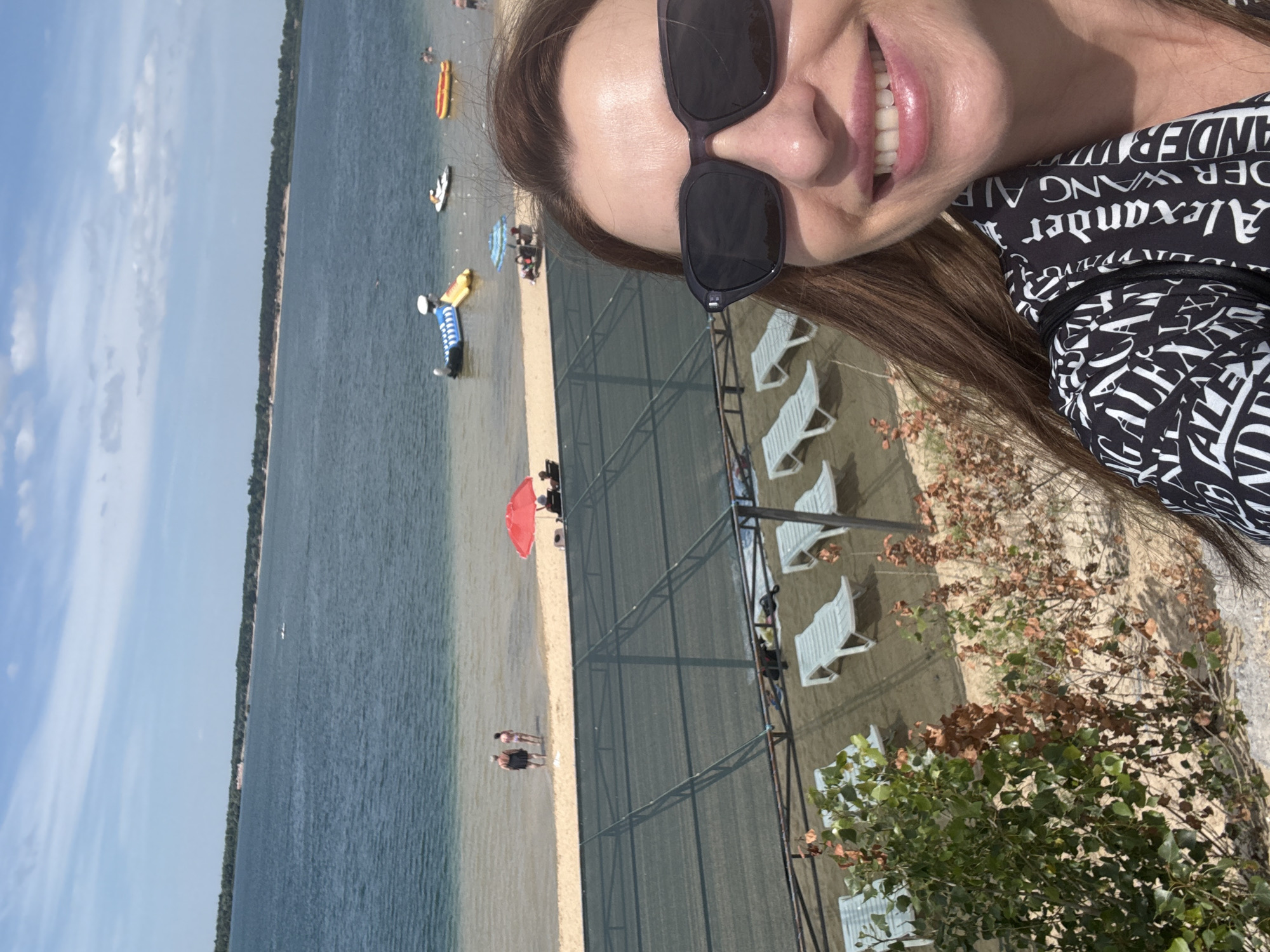 This is a selfie taken by a woman on a sunny day at a beach or waterfront area. The woman is wearing sunglasses and a black and white patterned top. She is smiling, with her teeth visible. In the background, there is a body of water, a sandy beach with lounge chairs, and some vegetation. There are also a few people visible on the beach and in the water, some with inflatable toys. The scene suggests a recreational outing. The text visible on the woman's top says "Alexander Wang."