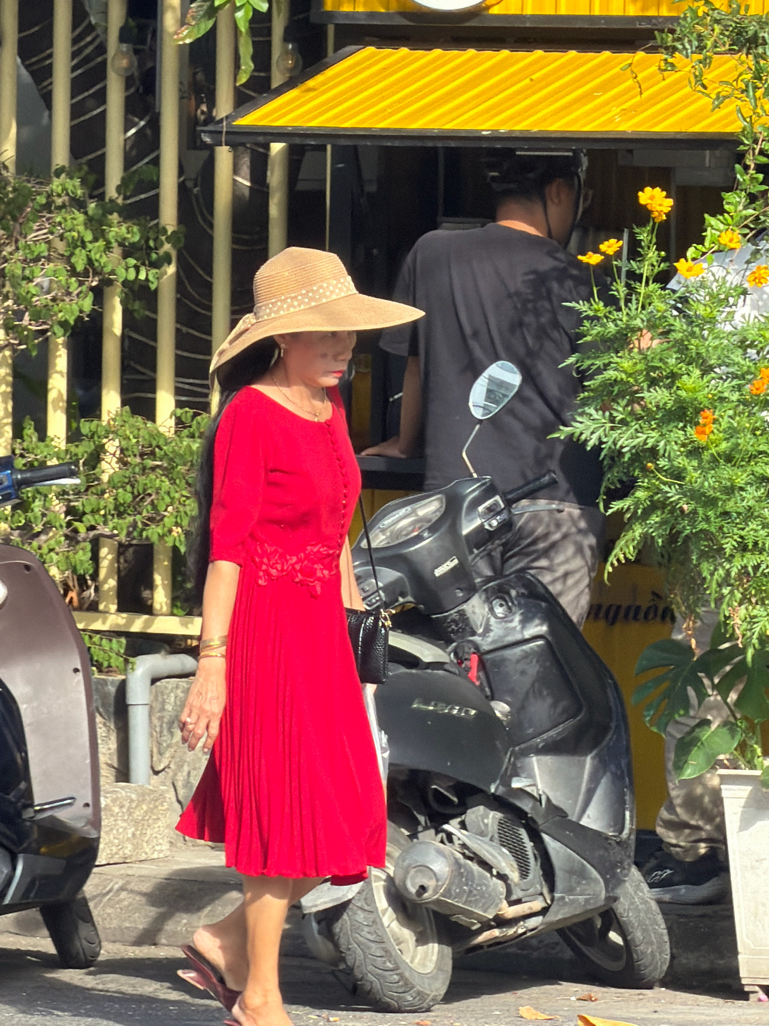 A woman wearing a wide-brimmed straw hat and a vibrant red dress with pleats and floral embellishments walks past a motorcycle in a city street. She has long dark hair and is adorned with several gold bracelets and a necklace. Her right hand is held near her mouth, possibly holding a cigarette. In the background, a person in a black t-shirt is visible near a yellow awning. The scene appears to be outdoors, with greenery and a plant in a pot to the right of the frame. The overall setting suggests a bustling urban environment, possibly in Nha Trang, Vietnam, given the visual cues often associated with such locations.