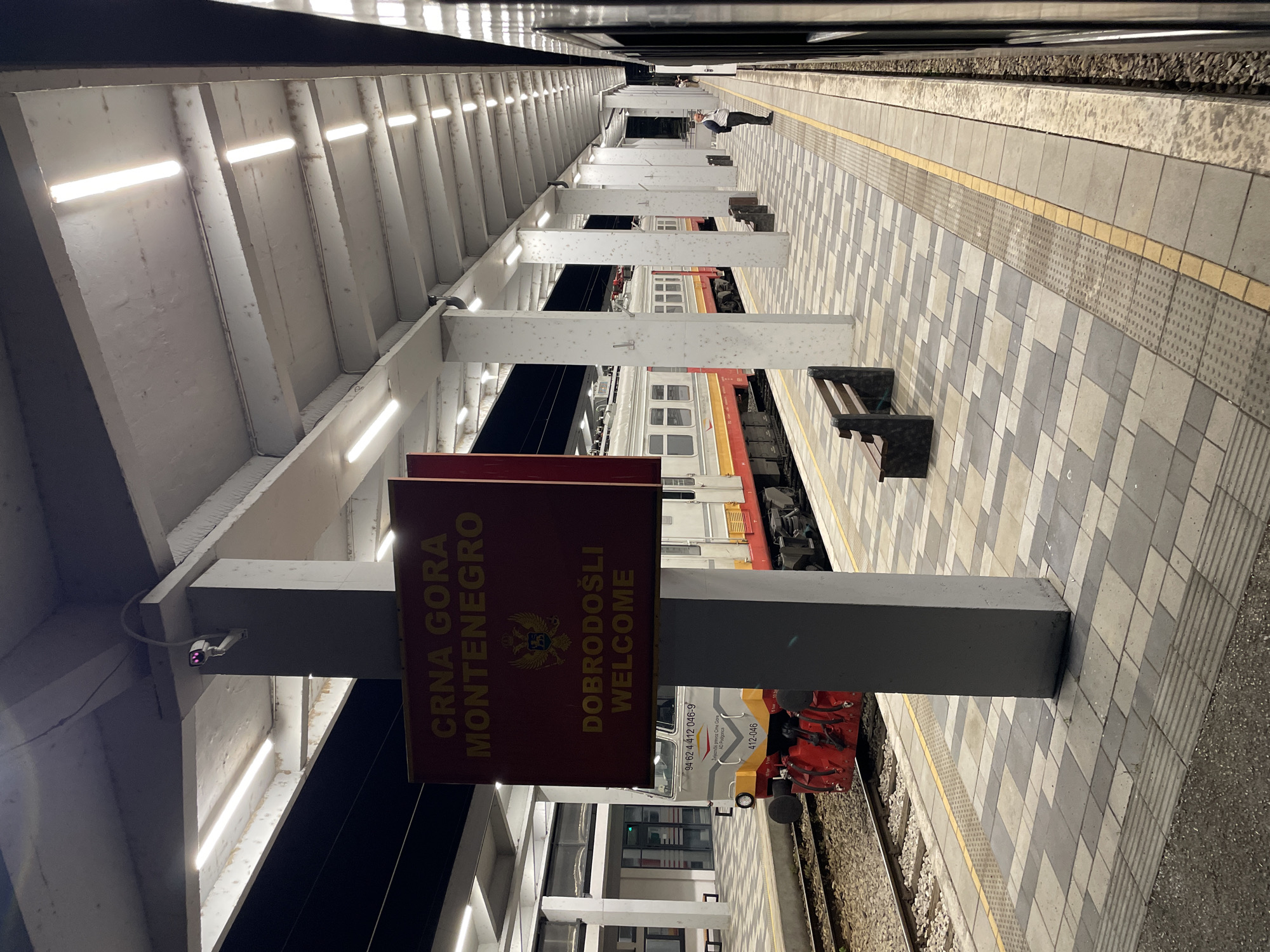 The image depicts a train station platform at night. A sign with the text "CRNA GORA MONTENEGRO DOBRODOŠLI WELCOME" and the coat of arms of Montenegro is prominently displayed. A train is visible on the tracks to the left, with its front engine painted in red, white, and yellow. The platform is paved with square tiles in shades of gray and white, with a yellow tactile warning strip along the edge. Several concrete pillars support an overhead structure with fluorescent lighting. A single wooden bench is placed on the platform. In the background, a person in a dark jacket and light-colored vest is standing near the edge of the platform, looking towards the train. The overall environment suggests a functional and well-lit transportation hub. The location is identified as Nedakusi, Montenegro.