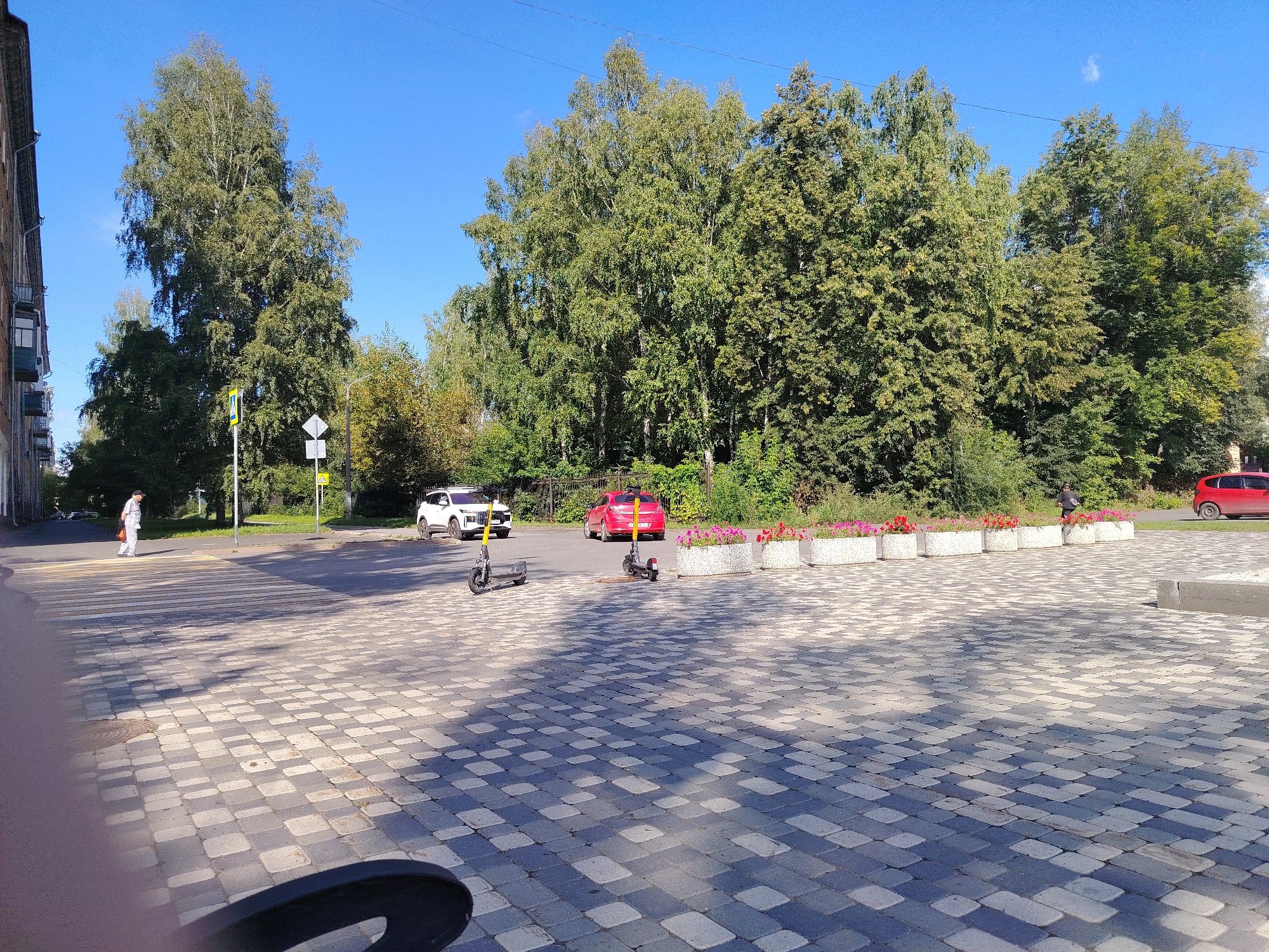 The image depicts a street scene in Kemerovo, Russia, on a clear, sunny day. In the foreground, a paved area with a patterned grey and white brick surface is visible, with shadows cast by an unseen object. Two electric scooters are parked on this area. Further back, a pedestrian crossing is marked on an asphalt road. A person dressed in light-colored clothing is walking across the pedestrian crossing. To the left of the road, a building with balconies is partially visible, and a tall birch tree with green foliage stands behind it. Across the road, a white SUV and a red hatchback are parked. In the background, a dense line of green trees forms the horizon, and a clear blue sky occupies the upper portion of the image. Flower boxes filled with pink and red flowers are positioned along the edge of the road between the parked cars and the trees. A power line is visible stretching across the sky.