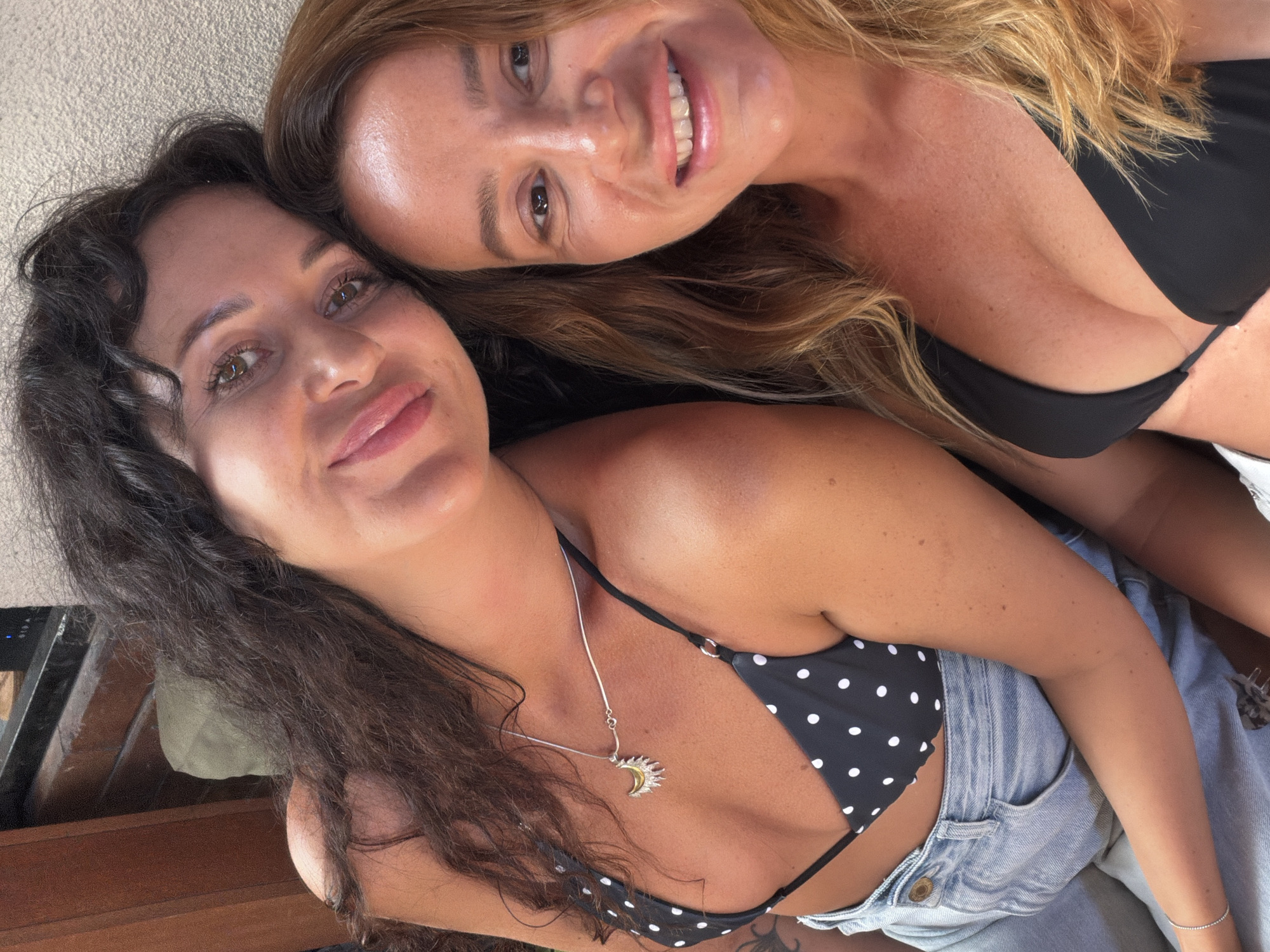 Two women are pictured in a close-up, selfie-style shot. The woman on the left has dark, curly hair and is wearing a black and white polka dot bikini top and light wash denim shorts. She has a necklace with a crescent moon pendant. The woman on the right has blonde, wavy hair and is wearing a black bikini top. Both women are smiling and looking towards the camera. The background is blurred, but appears to be an outdoor setting, possibly a patio or poolside area. The lighting suggests it is daytime.