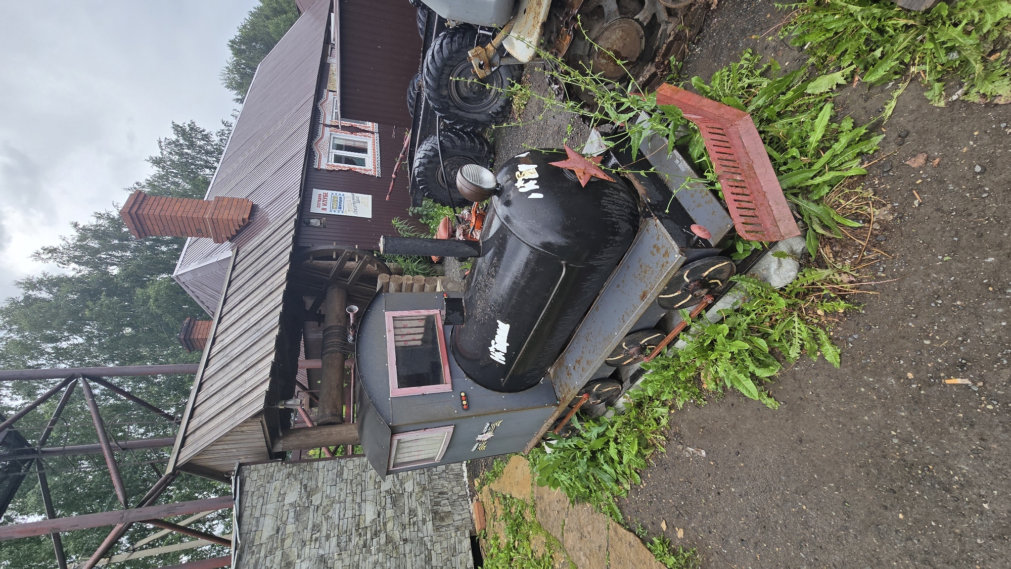 A small, black, metal, toy train sits on a track outside a building. The train has a red star on its front and white writing on its side. It is surrounded by green weeds and a dirt path. In the background, there is a building with a red roof and brick chimney, as well as a metal structure and trees. The sky is overcast. This scene is located in Bol'shoi Mashlyak, Russia.