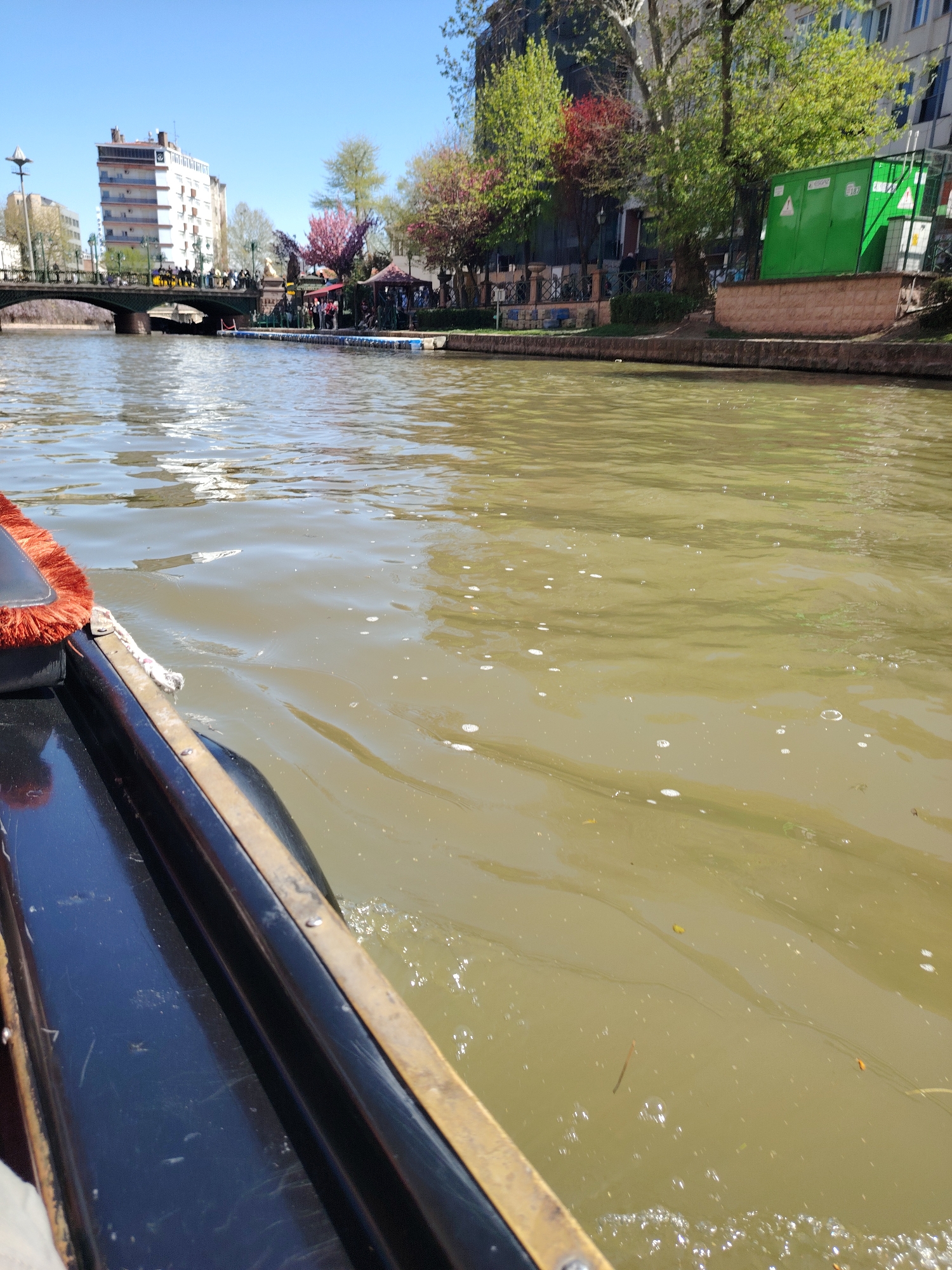 The photograph depicts a river scene in Eskisehir, Turkey, as viewed from a moving boat. The water is murky, with light ripples and visible bubbles. The boat's black side is prominent in the foreground, detailed with tarnished brass or gold-colored trim and a portion of the red-fringed cushion. Across the river, there is a pedestrian bridge filled with people, a docking area with additional individuals, and a line of trees with varying shades of green and red. Buildings are visible beyond the trees. A green utility box sits on the edge of the riverbank. The sky is clear and blue.