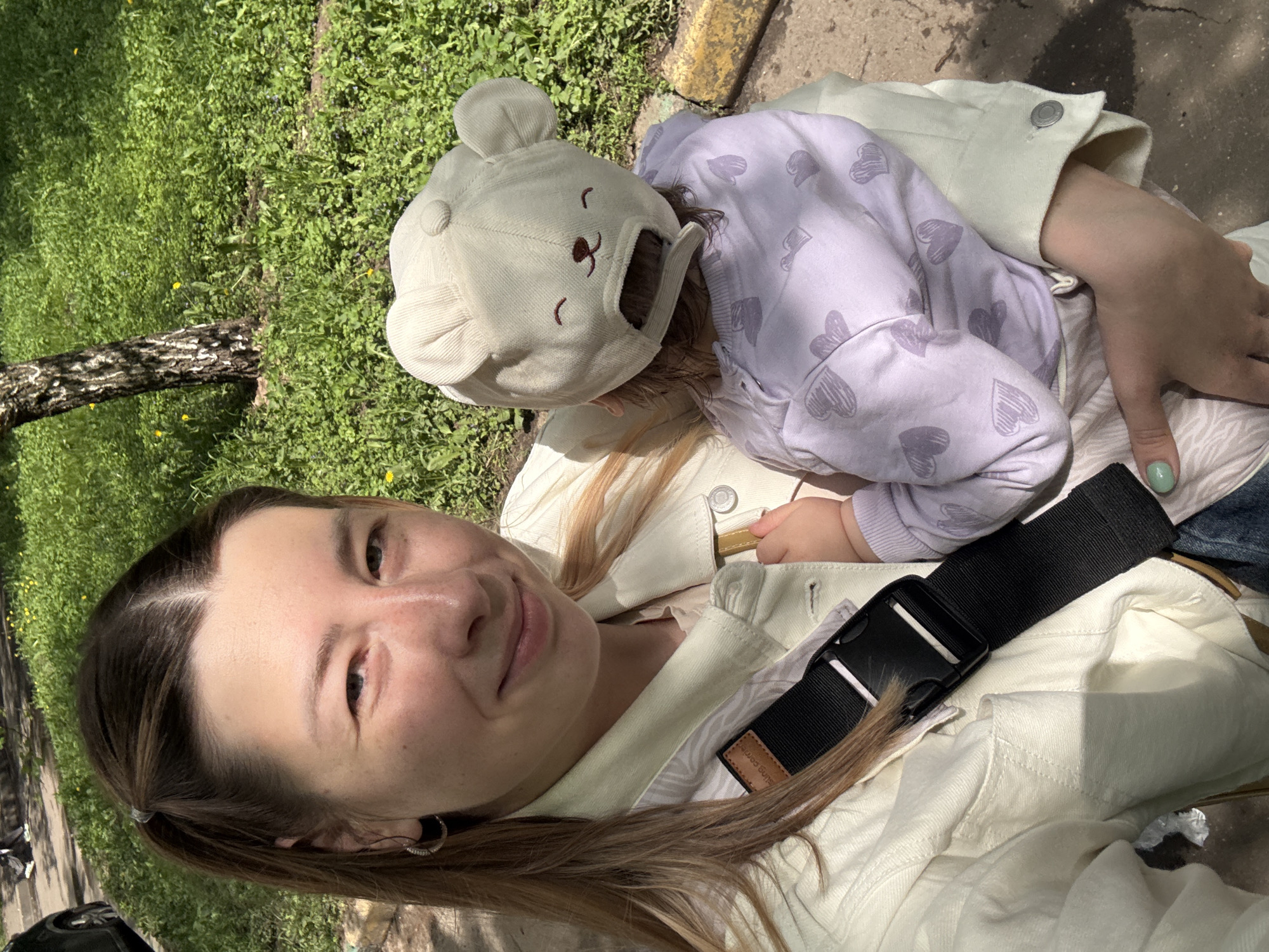 A woman holds a young child with a bear-shaped hat. The child wears a lavender shirt with grey hearts. The woman wears a light-colored jacket. A black strap crosses her chest. The scene appears to be outdoors in a grassy area with a tree trunk visible. Given the information, it is likely this photo was taken in a Moscow park in Russia.