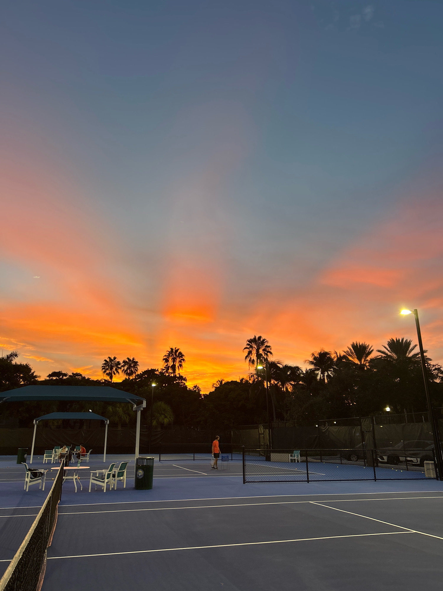 The image captures a sunset at a tennis court facility in Aventura, Florida. The sky is a gradient of colors, transitioning from a pale blue at the top to vibrant oranges and pinks closer to the horizon, with light rays emanating from behind a line of silhouetted palm trees. In the foreground, a bright blue tennis court with white lines is visible, with a net stretched across it. To the left, several chairs and a table are arranged under a shaded canopy. In the mid-ground, a person in an orange shirt is standing on the court. Further back, more tennis courts are visible, with fences, lights, and more palm trees and foliage. A single street lamp on the right illuminates a portion of the court. The overall scene suggests an evening setting at an outdoor recreational facility.