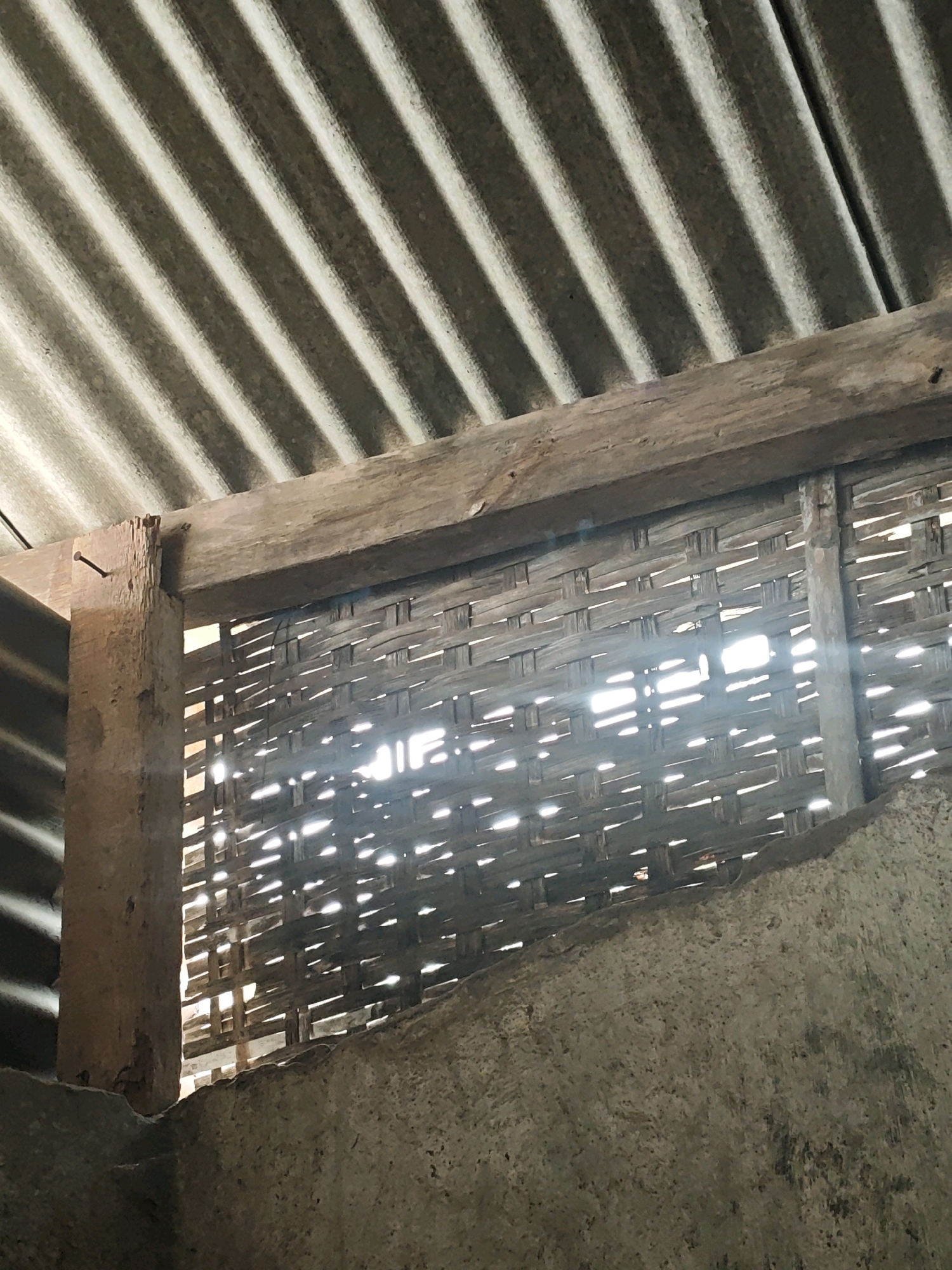 In Pagu, Indonesia, this image depicts the interior of a rustic structure with a corrugated tin roof angled overhead. A weathered wooden beam spans horizontally, supporting the roof and framing a woven bamboo wall. Sunlight streams through the gaps in the bamboo, creating bright streaks and casting shadows. On the left, a vertical wooden post, rough and partially splintered, has a nail protruding from it. Below the woven wall, a rough concrete structure forms the lower boundary of the scene. The overall impression is one of a simple, perhaps rural, construction. There are no people or animals visible in the image.