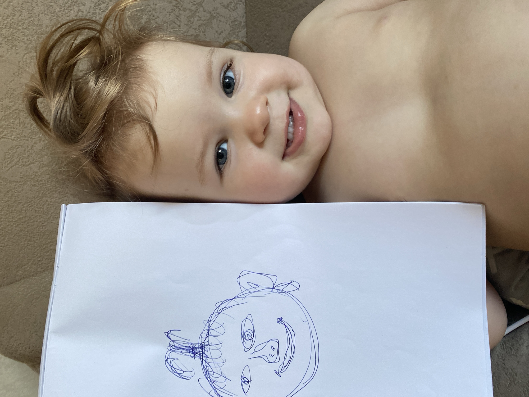 An overhead shot shows a smiling baby or toddler lying on a textured, light brown surface, likely a couch or bed, with a white sheet of paper held in front of them. The baby's head is turned slightly to the right, and they are looking directly at the camera with wide, bright blue eyes. They have fair skin, plump cheeks, and a happy, open-mouthed smile, revealing a few tiny teeth. Their light brown or blonde, curly hair is visible around their head, with some strands appearing to be tied up or naturally styled in a playful way.
The paper, which appears to be part of a sketchbook or pad, displays a child's drawing in blue ink. The drawing depicts a stylized face, possibly a pig or a cartoon character, featuring large, round eyes with spirals inside, a snout-like nose with nostrils, and a wide, upturned smile. Swirly lines suggest hair or ears around the head of the drawn figure. The baby's bare chest and shoulder are visible, suggesting they are lightly dressed. The overall impression is one of childlike joy and creativity in a comfortable, indoor setting during daylight hours. No visible text is present in the image.