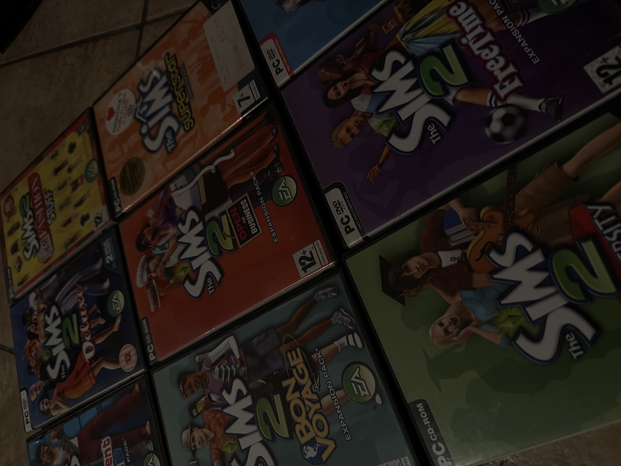 The image displays a collection of Sims 2 PC game cases arranged on a tiled surface. The games are stacked and slightly overlapping, creating a layered effect. The lighting is dim, with some cases catching more light than others, causing reflections and obscuring some details.
The visible game titles include:
* The Sims 2 Deluxe
* The Sims 2 Fashion Stuff
* The Sims 2 Superstar
* The Sims 2 Open for Business
* The Sims 2 Bon Voyage
* The Sims 2 FreeTime
* The Sims 2 University
The image does not contain any information to suggest a location in Sharm El Sheikh, Egypt, or any other specific geographic location. The focus is solely on the game cases and their arrangement.