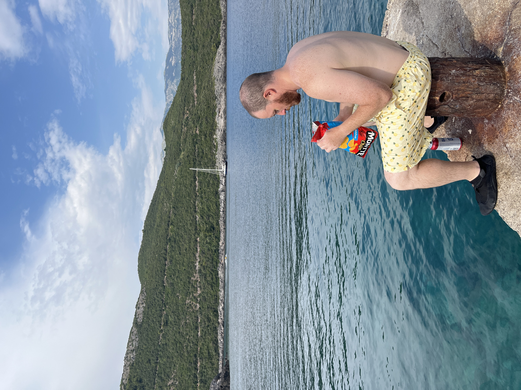 A shirtless man with a beard sits on a rustic wooden stump on a concrete pier, holding a bag of Monte Chips. He is wearing light yellow shorts with a pineapple pattern and black slip-on shoes. To his right, a red and silver can rests on the pier. The sea in front of him is a vibrant turquoise, with gentle ripples reflecting the blue sky and scattered white clouds. In the distance, lush green hills rise from the coastline, dotted with trees. A white sailboat is anchored in the bay. The overall scene suggests a relaxed moment by the water in a scenic, coastal environment, likely near Dubraljevina, Montenegro, given the picturesque mountainous landscape and clear blue sea.