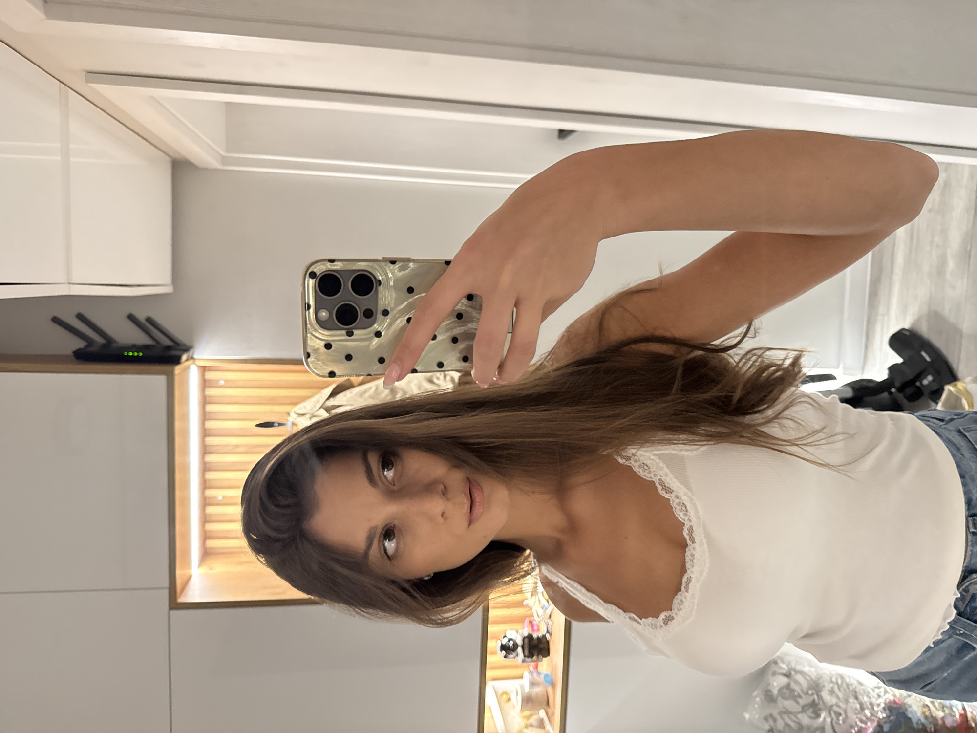 A woman is taking a selfie in front of a mirror. She is holding a phone with a dotted case. She is wearing a white tank top with lace trim and blue jeans. Her hair is long and brown. She is looking at the camera. Behind her is a white cabinet with a black router on top and a lit display case. There is also clothing and shoes behind her. There is no evidence to suggest a location in Burgas, Bulgaria.