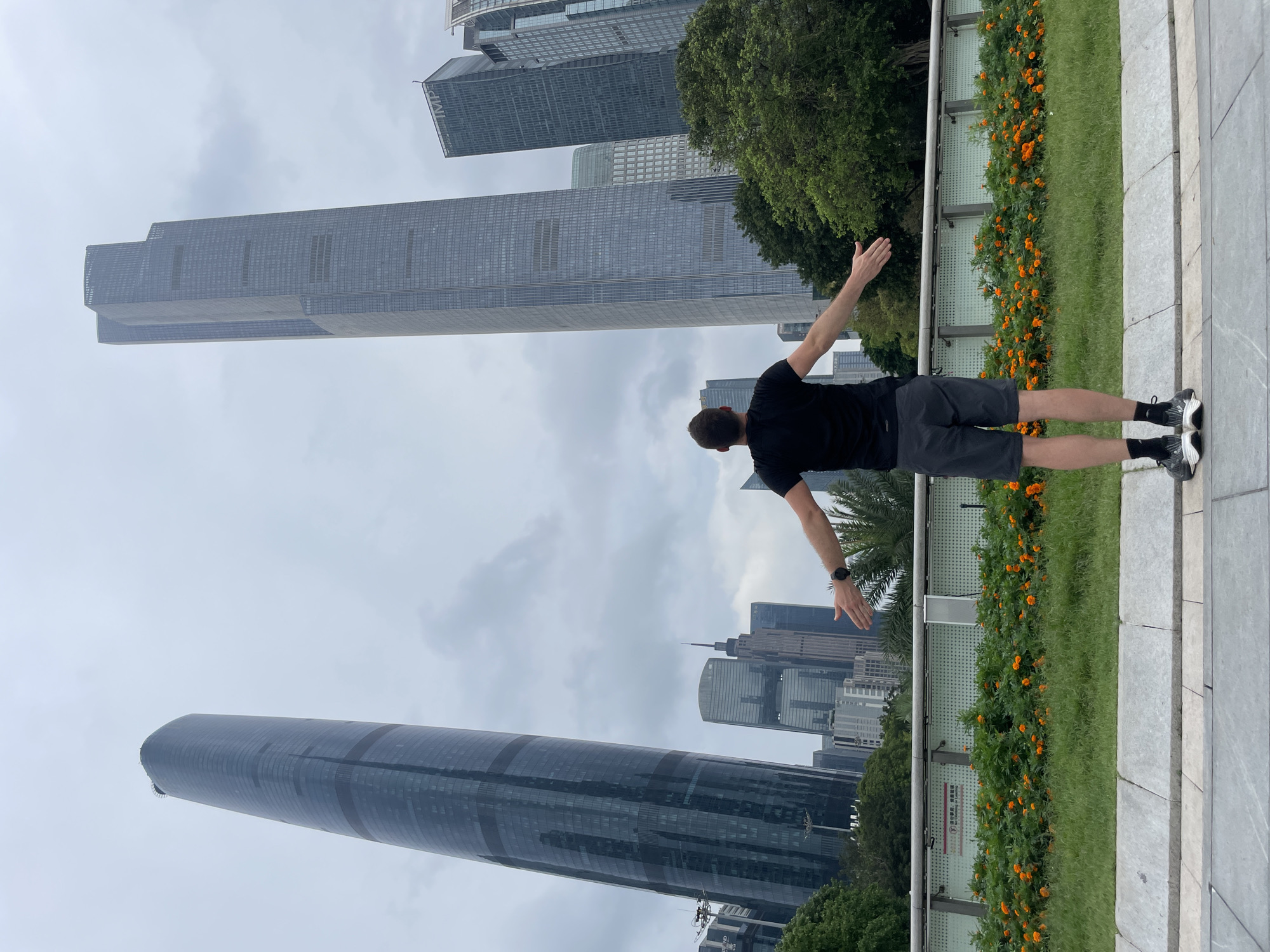A man stands with his arms outstretched in the city of Tianhe District, China. The man is wearing a black t-shirt and gray shorts. He is standing on a sidewalk next to a flower bed with orange flowers. In the background are several tall skyscrapers. The sky is overcast.