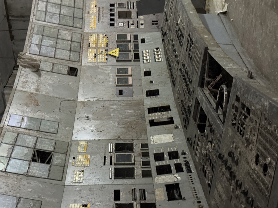 This image depicts an abandoned and severely deteriorated control room, characteristic of an industrial facility, specifically resembling the control rooms found within the Chernobyl Nuclear Power Plant, located in the Chernobyl Exclusion Zone, north of Kyiv, Ukraine. No people are present.
The foreground features a large, curved control console, dark gray and heavily encrusted with grime and dust. Numerous square and circular cutouts reveal missing buttons, switches, and panels, leaving empty cavities and exposed wiring. Debris, including what appears to be organic material and discarded items, is visible within these hollowed-out sections.
Behind the console, a multi-tiered wall of vertical panels, lighter gray and extensively corroded, displays peeling paint and heavy staining. The lower and middle tiers exhibit empty rectangular slots where instruments once resided, alongside faded yellow-on-black labels and rows of small, dark, possibly non-functional indicators. A prominent yellow triangular radiation hazard symbol with a black trefoil is affixed to a panel on the middle tier, slightly right of center. The upper tier consists of multiple shattered or missing display windows, revealing dark interiors and underlying structural elements. Loose wires hang from various panels, and a piece of light-colored fabric or plastic dangles from the ceiling near the top center.
The scene portrays significant neglect and advanced decay, indicative of prolonged abandonment in an unpowered, dimly lit environment.