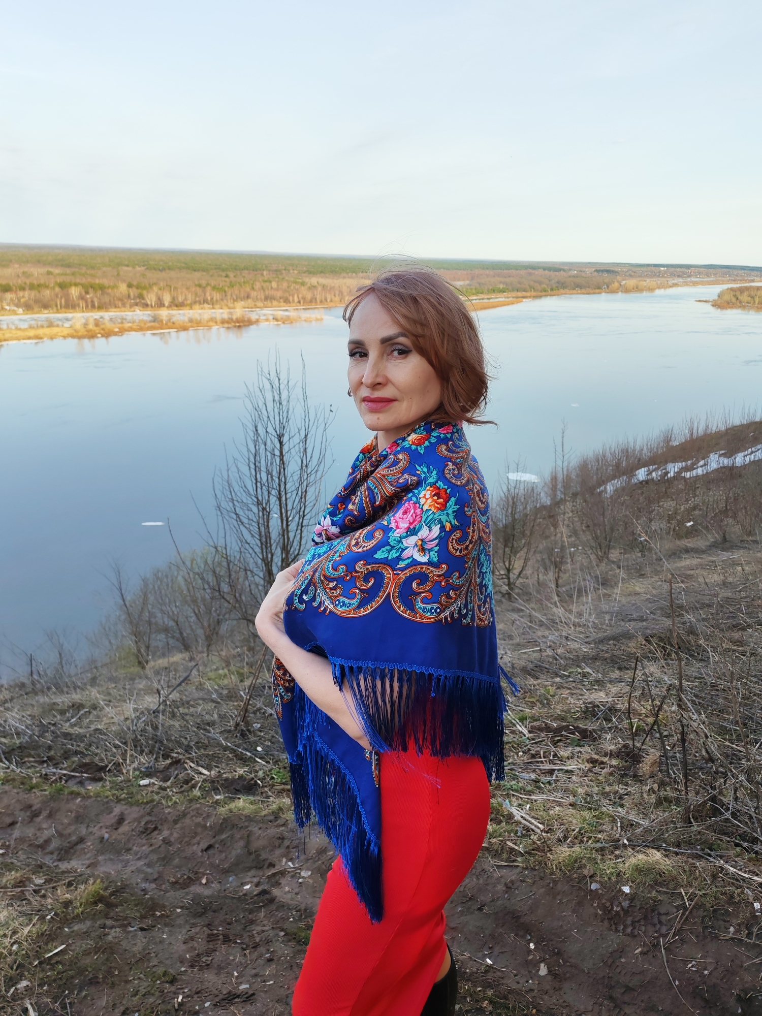A woman stands on a high riverbank in Sarapul, Russia. She is dressed in red trousers and a blue floral shawl with fringe. The Volga river stretches out behind her in the background. The riverbanks are a mix of trees and shrubs. The sky is overcast.