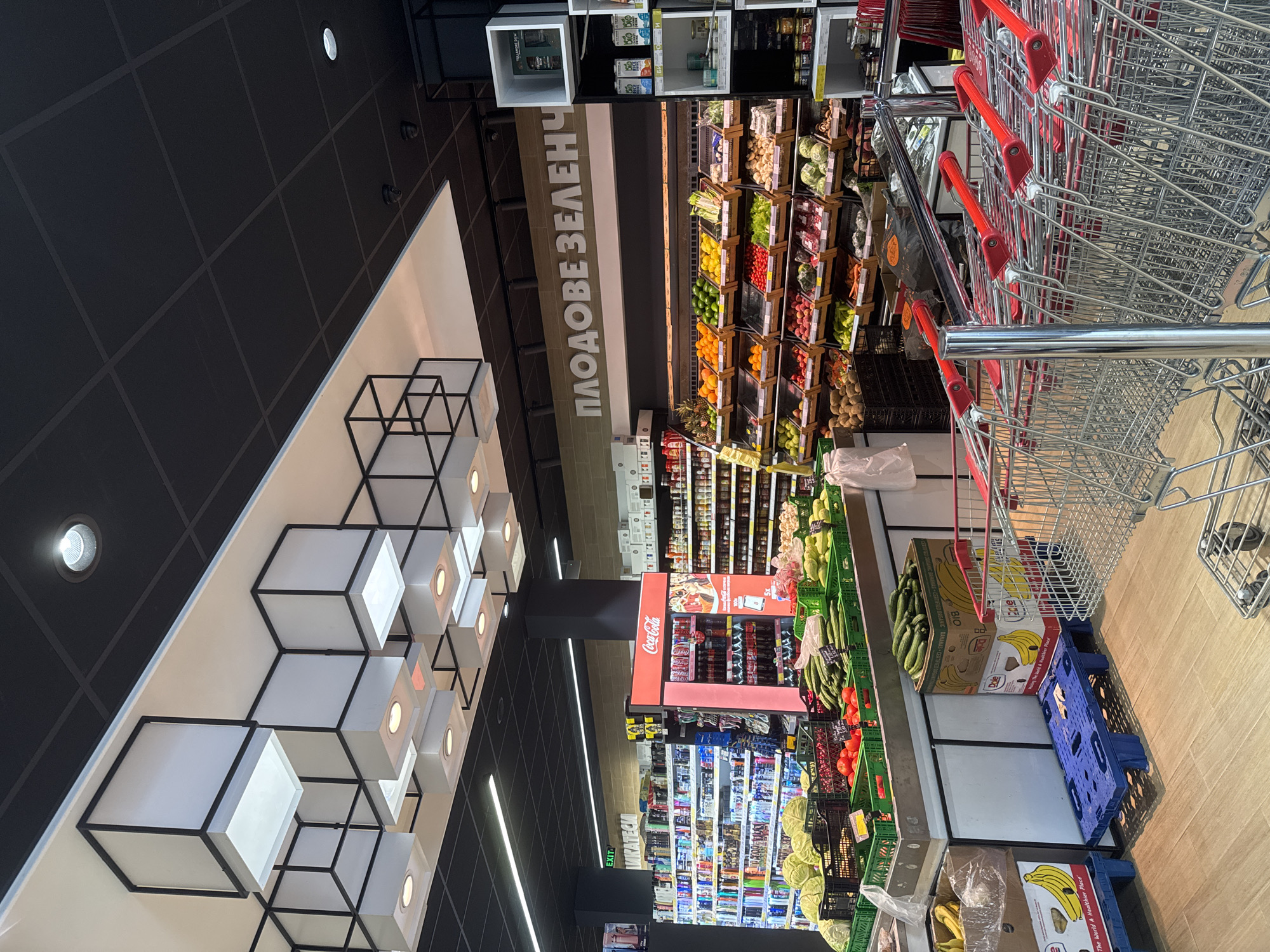 The image depicts the interior of a supermarket in Varna, Bulgaria, specifically the produce section. The ceiling features modern, cube-shaped lighting fixtures and a black grid pattern. In the foreground, a display of fresh produce is visible, including cabbages, tomatoes, cucumbers, and various fruits arranged in wooden crates and green plastic containers. Banana boxes are also stacked nearby. Multiple rows of empty shopping carts with red handles are lined up, suggesting a less crowded shopping period. In the background, shelves stocked with beverages and other grocery items can be seen, along with a prominent Coca-Cola branded refrigerator. A sign with Cyrillic text, "ПЛОДОВЕ ЗЕЛЕНЧУЦИ" (Fruits Vegetables), is mounted above the produce display. The overall environment is clean and well-lit, typical of a retail grocery store. No people are visible in the image.