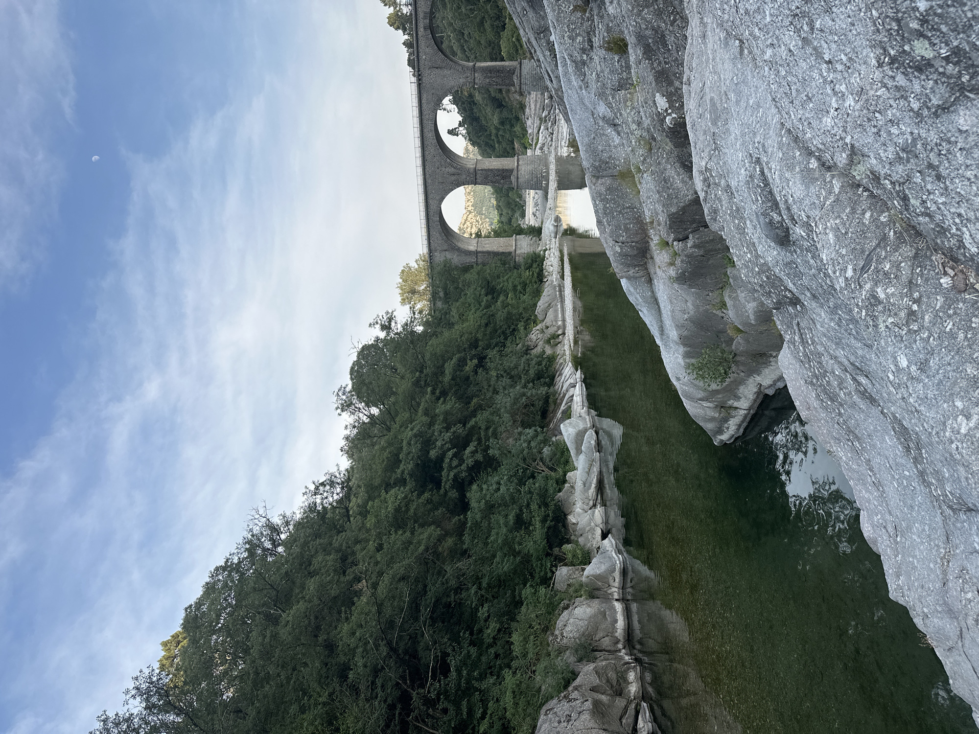 This media file captures a scenic view of a stone viaduct with multiple arches, spanning across a narrow gorge. The viaduct is situated amidst lush green trees and vegetation, suggesting a natural, possibly mountainous setting. Below the viaduct, a calm body of water reflects the surrounding landscape and the stone structures above. The water's surface is partly broken by ripples and reflections of trees and sky.
On the right side of the image, a large, weathered rock formation dominates the foreground, adding a sense of scale and natural grandeur to the scene. The sky above is partly cloudy, with some wispy white clouds against a light blue backdrop. A small crescent moon is faintly visible in the upper left portion of the sky.
The lighting suggests it is daytime, likely late afternoon or early morning, given the soft light and the presence of the moon. The overall mood is tranquil and serene, highlighting the harmonious coexistence of man-made architecture and the natural environment. There are no people visible in the image, and no text is present. The scene appears to be a peaceful, picturesque location, possibly a park or a natural reserve, inviting contemplation and appreciation of its beauty.