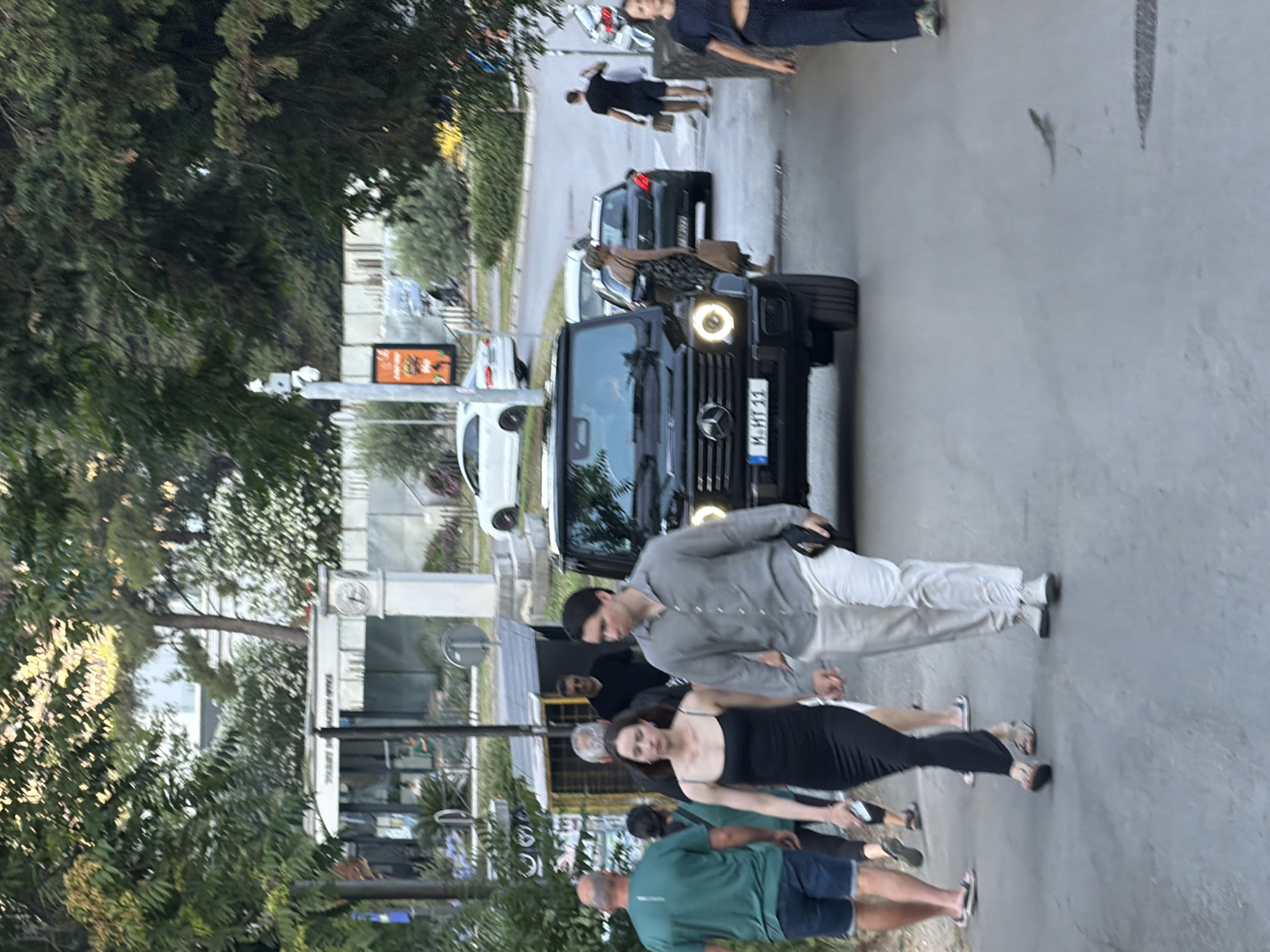 In Budva, Montenegro, a black Mercedes-Benz G-Class SUV is parked on a street, with its headlights illuminated. A young couple, holding hands, walks in front of the vehicle. The woman wears a black dress, and the man is in a light-colored shirt and pants. Behind them, a man in a green shirt and shorts walks away from the camera, and other pedestrians are visible in the background, including a man in a black shirt and shorts, and a woman in a dark top and pants. The scene appears to be a commercial area with buildings and trees lining the street. A clock tower is visible in the distance.