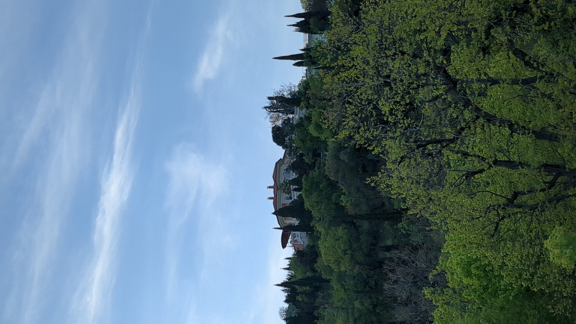 The image depicts a building on a hill surrounded by trees, with a blue sky featuring wispy clouds in the background. The location is Unknown, Unknown. The building appears to be multi-storied, with a red roof. Green trees dominate the foreground and midground.