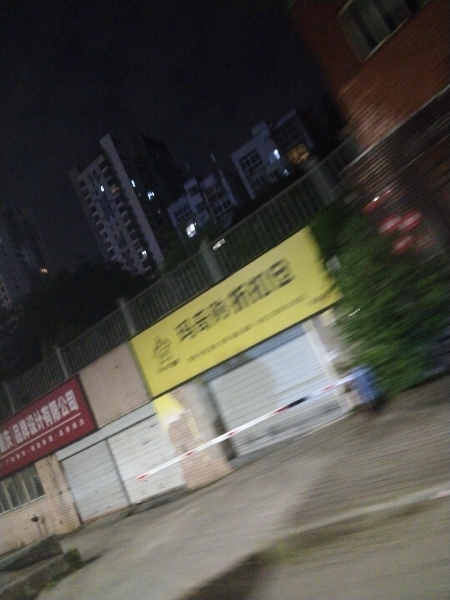 The image depicts a street scene at night in what appears to be Jiulongpo District, China, based on the presence of Chinese signage. The scene includes a row of closed storefronts with metal roll-up doors. Above the storefronts are signs; one is red with white Chinese characters, another is yellow with black Chinese characters. A red and white barrier tape stretches across the front of the storefronts. In the background, there are residential buildings. The lighting is dim and the image appears to be blurred due to motion. No people are clearly visible. Vegetation is present to the right of the frame. The ground is paved and appears to be a sidewalk or street.