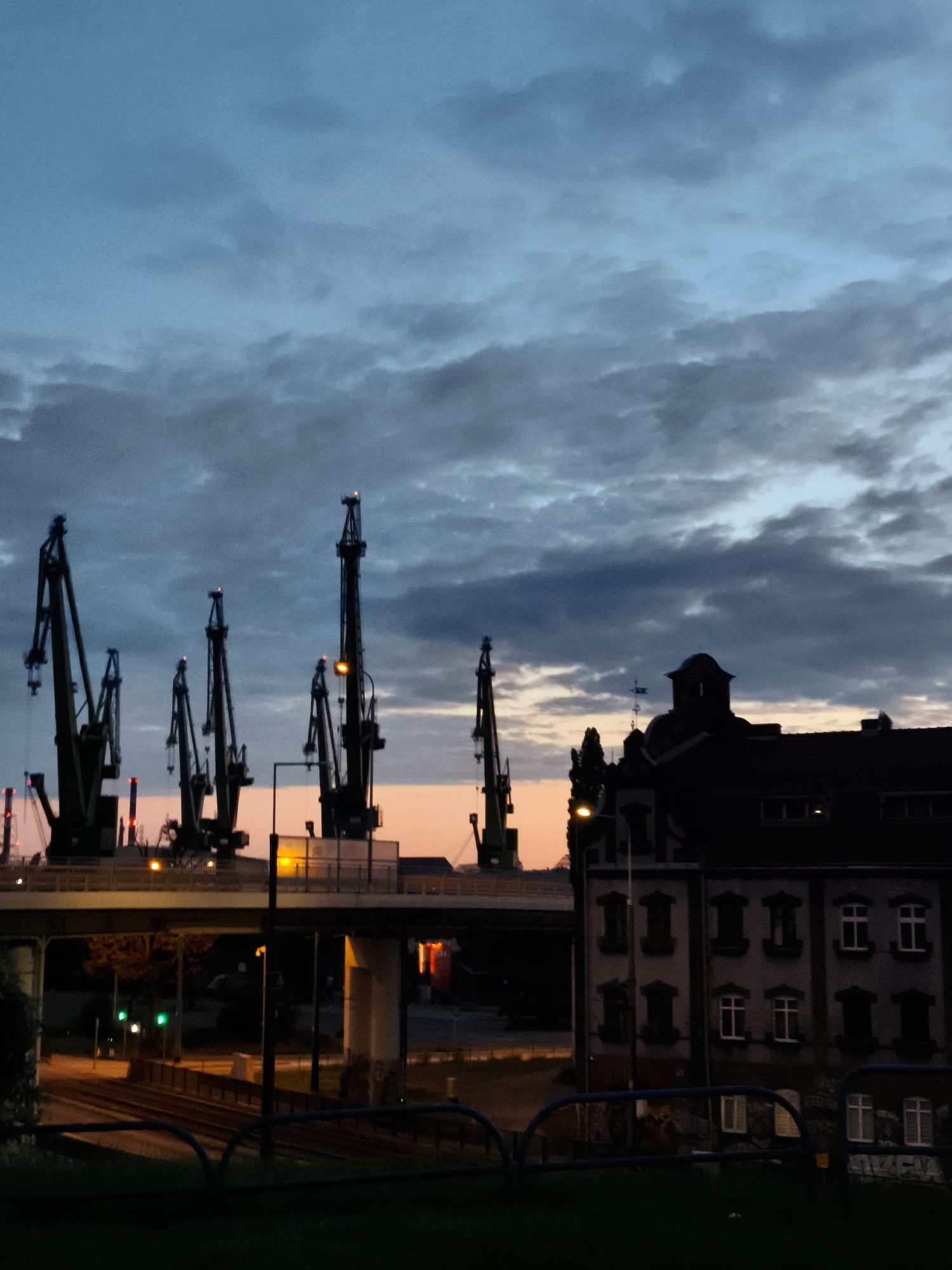 This is a vertical, high-angle, outdoor shot of the Gdańsk Shipyard in Gdańsk, Poland, at dusk. The sky is a gradient of deep blue and gray, with darker clouds obscuring a faint orange glow near the horizon. Silhouetted against the sky are several large industrial cranes, their structures dark and imposing. To the right, a multi-story building with a distinct domed roof also appears in silhouette. In the foreground, a dark, grassy area is bordered by a blue metal fence. A roadway with train tracks and traffic lights, some illuminated green, runs beneath an elevated structure that supports some of the cranes. The lighting suggests a low-light condition, possibly after sunset.