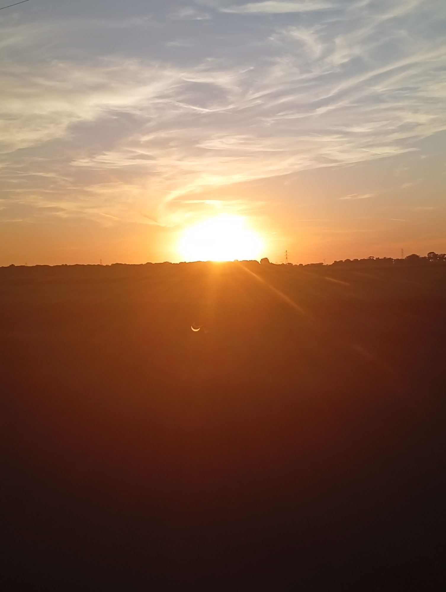 This is a photograph of a sunset over a rural landscape. The sun is low on the horizon, casting a warm orange glow across the sky. Wispy clouds are scattered across the sky, and the landscape below is silhouetted against the bright sky. A crescent-shaped object is visible in the lower part of the image, slightly to the left of the center, which appears to be a lunar eclipse. The terrain is relatively flat, with some distant trees and structures visible along the horizon. The image is taken from a low angle, looking out towards the horizon.