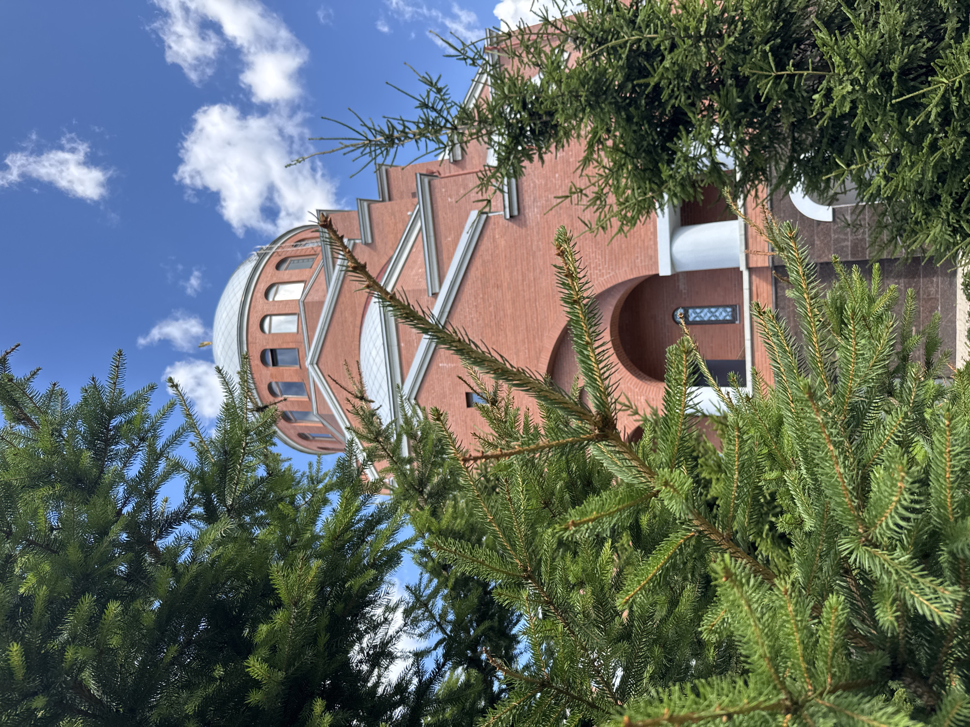 The image shows the Church of the Resurrection of Christ in Sinyavino, Russia, partially obscured by lush green pine trees. The church is constructed of red brick and features a prominent silver dome topped with a golden cross. Its architecture includes arched windows and white columns. The sky is a vibrant blue, dotted with fluffy white clouds. The foreground is dominated by the dense foliage of the evergreen trees, creating a layered effect with the church in the background. The lighting suggests it is daytime, likely sunny.