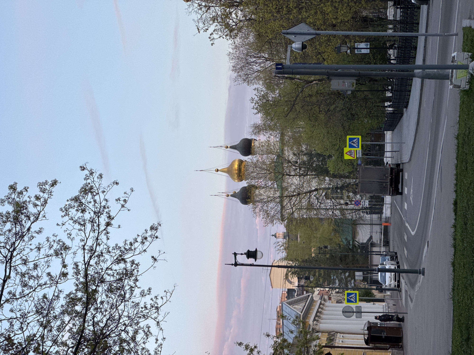 The image depicts a street scene in Moscow, Russia. The photograph is taken from an angle that shows three onion domes of a Russian Orthodox church in the distance, one gold and two dark grey. The sky is a mix of light blue and pink hues. The street is lined with trees. There is a pedestrian crossing sign visible on the sidewalk. The road has painted lines marking lanes. A vehicle is traveling down the street. A street light is visible on the left side of the frame and buildings line the street. A tree without leaves is featured in the foreground of the left side of the image.