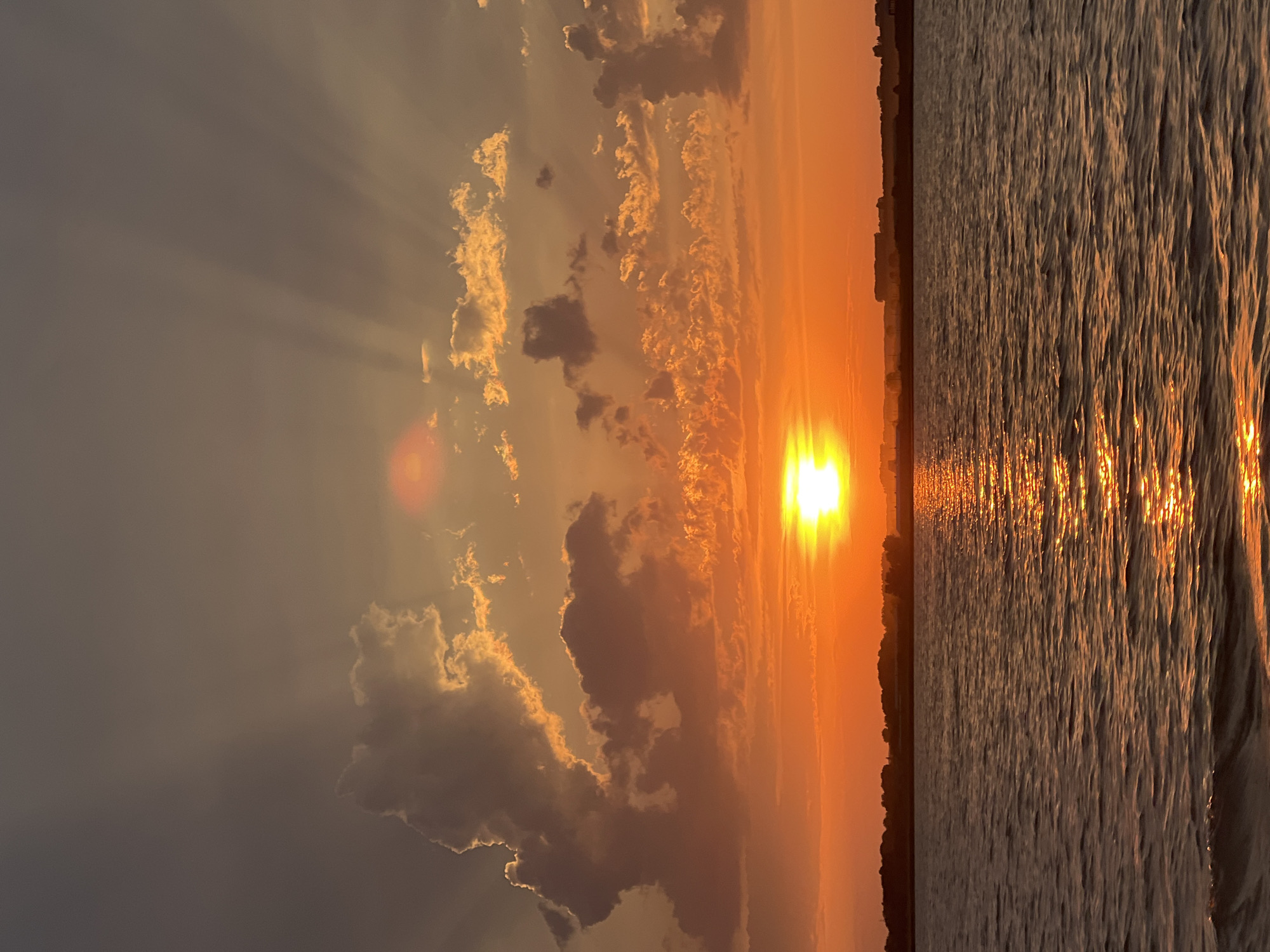 A vibrant sunset casts a fiery glow over the tranquil waters of Novopichugovo, Russia. The sun, a brilliant orb of orange, is positioned just above the horizon, its rays fanning out through scattered clouds. These clouds, painted in hues of orange and gray, are illuminated by the setting sun, creating dramatic shafts of light that pierce the sky. The water below reflects the sky's warm tones, with shimmering streaks of sunlight dancing on its surface. The distant shoreline is silhouetted against the bright sky, dotted with trees and buildings, suggesting a quiet, picturesque landscape. The overall scene is one of serene beauty, capturing the fleeting moments of twilight in a Russian setting.