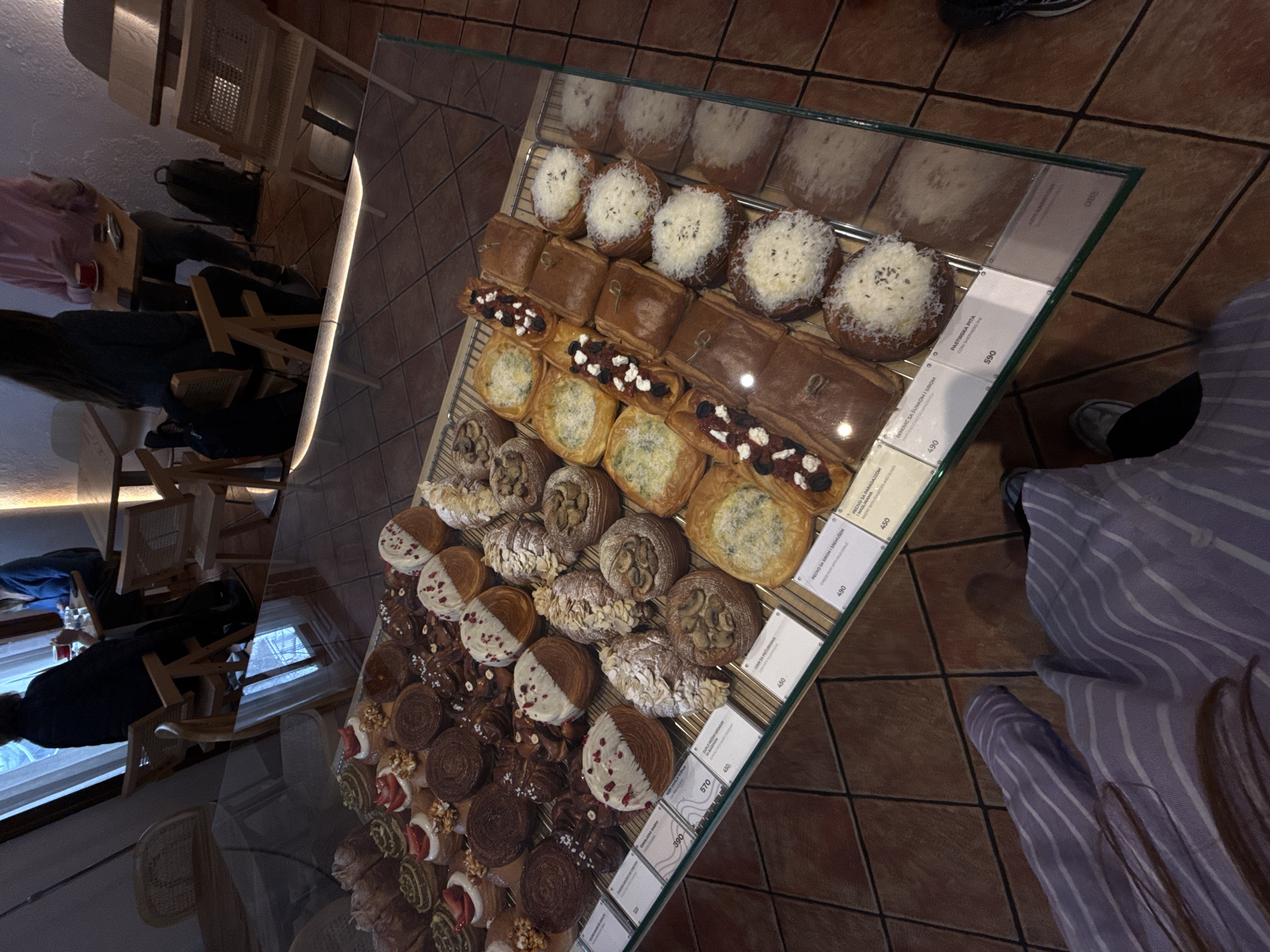 Captured from an overhead perspective, the photograph presents a clear display case stocked with an array of pastries. The pastries, ranging from cheese-filled delicacies to options with mushrooms, olives, and shepherd's pie, are accompanied by labels indicating their ingredients and prices in Serbian dinars. Notably visible are price tags for 570, 390, 450, 450, 490, and 590. Patrons can be seen observing the pastries from various angles. Situated indoors, the scene is set against a backdrop of tables and chairs. The photo appears to have been taken in Belgrade, Serbia, given the language on the price tags.