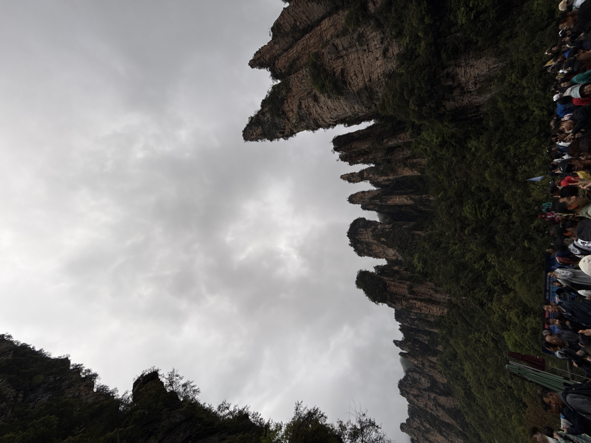 Looking up at rock formations in Luoguta, China, there are numerous people on the right side of the image, with the top of the rock formation visible, with a cloudy grey sky above. Rock formations are covered with green vegetation. Some trees are visible at the bottom left.