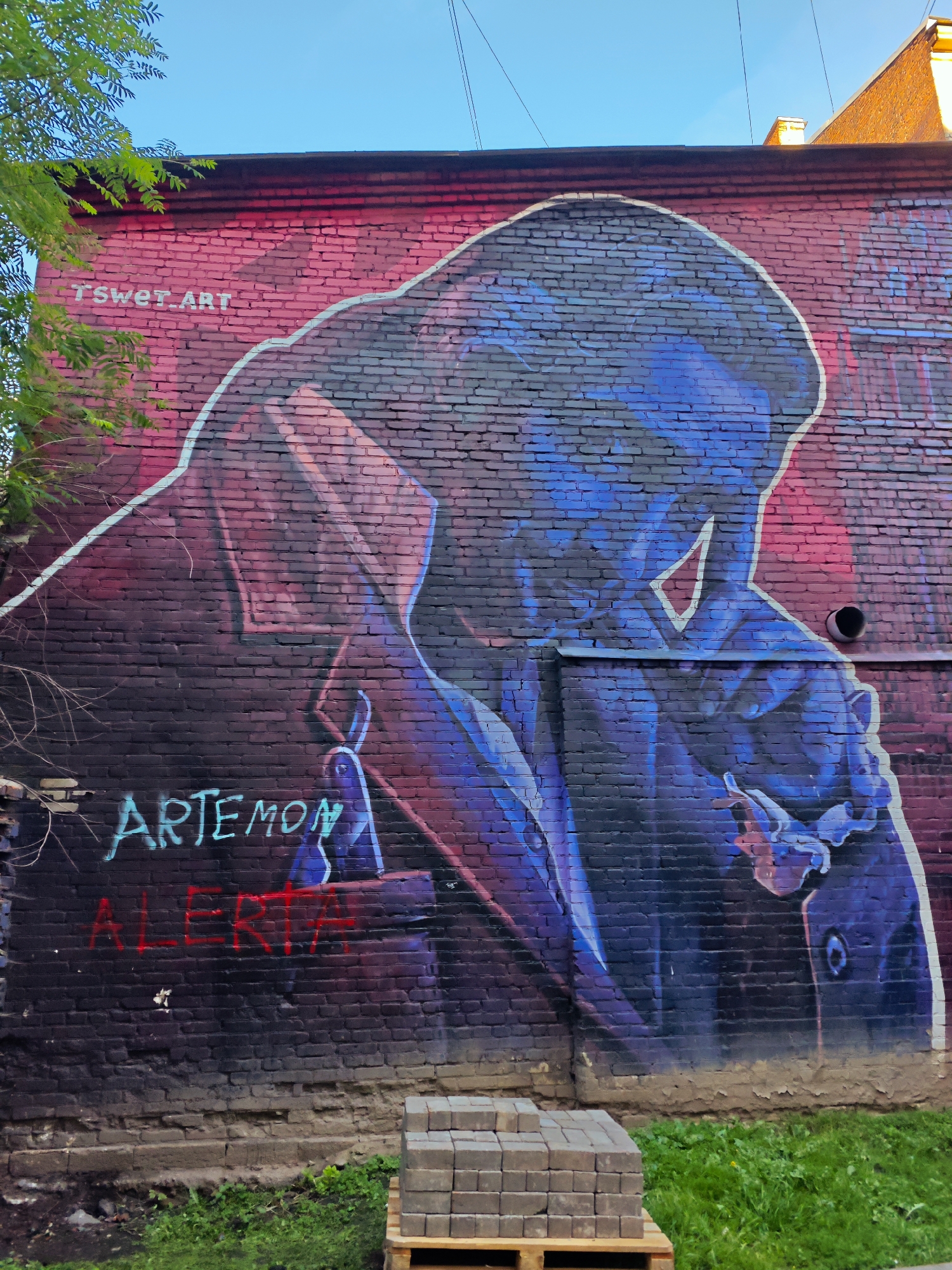 This image depicts a large graffiti mural on a brick wall in Saint Petersburg, Russia. The mural features a stylized portrait of a figure in a dark coat, rendered in shades of blue, purple, and red. The figure appears to be holding a cigarette. To the left of the figure, the text "TSwer.ART" is visible in white. Below the figure, the words "ARTEMON" are written in light blue, and "ALERTA" is written in red. The brick wall shows signs of wear and tear, with some crumbling at the base. In front of the wall, there is a patch of green grass and a pallet stacked with paving stones. The sky above is clear and blue.