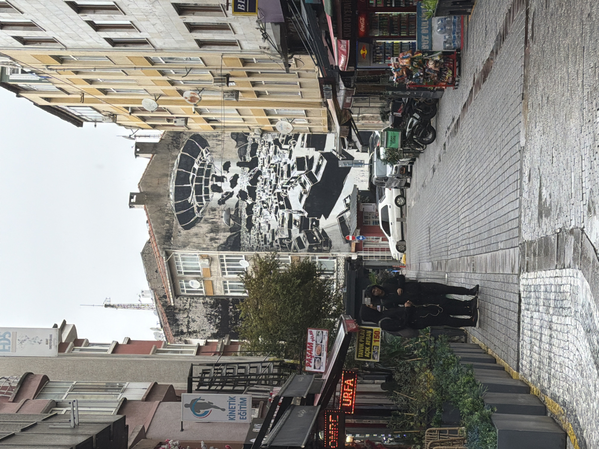 The image depicts a street scene in Kadıköy, Turkey. The street is paved with cobblestones, and the buildings lining the street are several stories high. On the left side of the street, there are shops with awnings and signs, including one for "KINEMTIC EGITIM" and another that reads "URFA". There are also planters with greenery. On the right side of the street, there are more shops, a refrigerator stocked with drinks, and a rack of snacks. A large mural depicting a futuristic, dystopian cityscape covers the side of a building in the background. In the middle of the street, there are several vehicles, including cars, vans, and a scooter. Three individuals, dressed in dark clothing, are standing on the sidewalk on the left side of the street, facing away from the camera. The sky is overcast, suggesting a cloudy or rainy day, with puddles visible on the street and sidewalk.