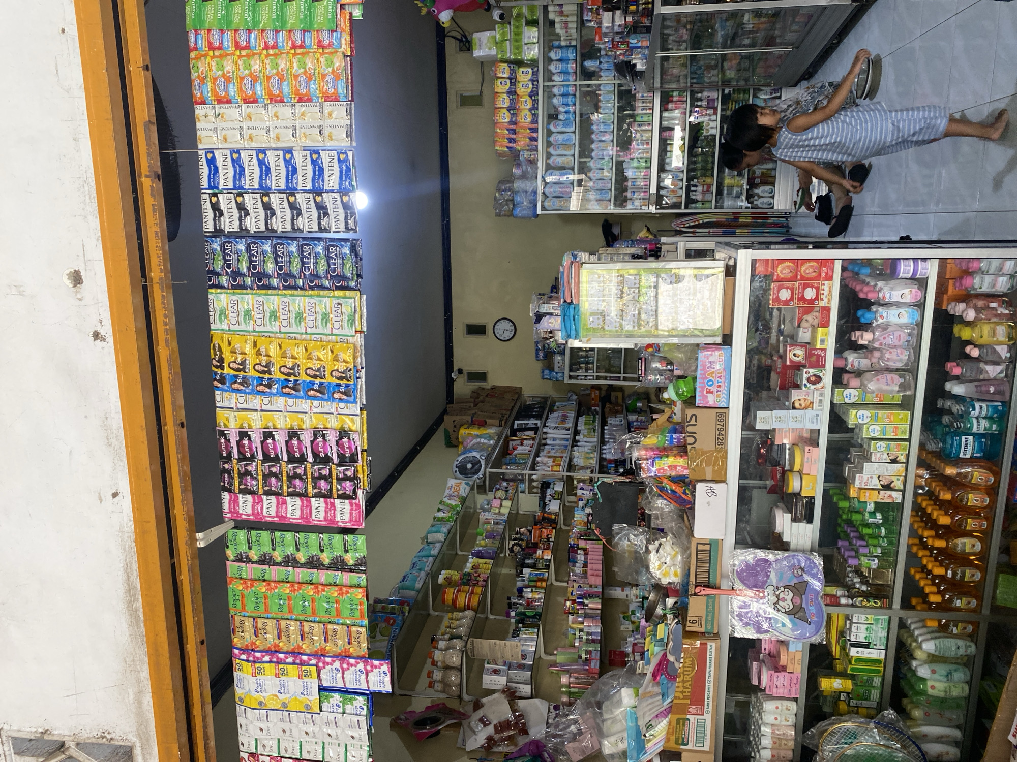 The image depicts the interior of a small retail store, likely a minimarket or a convenience store, located in Ta', Indonesia. The store is stocked with a variety of personal care products, cleaning supplies, and some household items.
Hanging prominently at the front of the store are rows of small sachets of shampoo and conditioner, brands like Pantene, Clear, Rejoice, and Sunsilk are visible. These are arranged in neat rows.
Inside the store, shelves are filled with numerous products. On the left, there are bottles of lotion, soap, and other personal care items. Further back, shelves are packed with more bottles, boxes, and containers, including what appear to be laundry detergents, dish soaps, and possibly cleaning sprays. Some items are still in their original packaging, while others are displayed more openly.
A glass-fronted display cabinet on the right side of the store showcases more bottled products, many of them clear or light-colored liquids. In the foreground, there are stacks of boxes and some loose items, including a fan, and what looks like a collection of colorful plastic toys or dispensers.
Two young children are present in the store. One child, a girl wearing a blue and white striped outfit, stands near the center of the image, looking to the right and holding a small black object. She is barefoot. Another child, partially visible in the background, is seated on the floor.
The store appears to be well-lit, with overhead lighting visible. The walls are painted a pale yellow or cream color, and the ceiling is dark. A clock is mounted on the wall in the background. The floor is tiled. The overall impression is of a functional, albeit somewhat cluttered, retail space serving a local community.