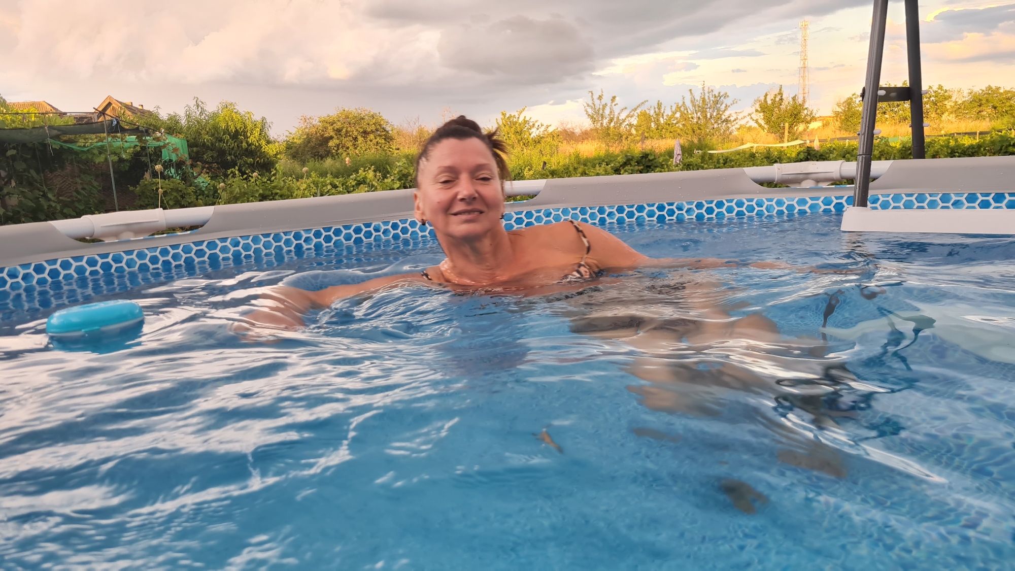 The image depicts a woman swimming in an above-ground pool on a cloudy day, with a backdrop of trees and houses suggesting a residential outdoor setting. The woman, with her hair pulled up, is wearing a patterned bikini top and appears to be smiling as she looks towards the camera. The pool is filled with blue water, and a blue float is visible on the left side of the frame. The pool structure has a grey frame and a blue hexagonal pattern along the upper edge. The background shows greenery, trees, and some buildings, with a cell tower visible in the distance. The lighting suggests it is either late afternoon or early morning. The location is identified as Salhany, Ukraine.
