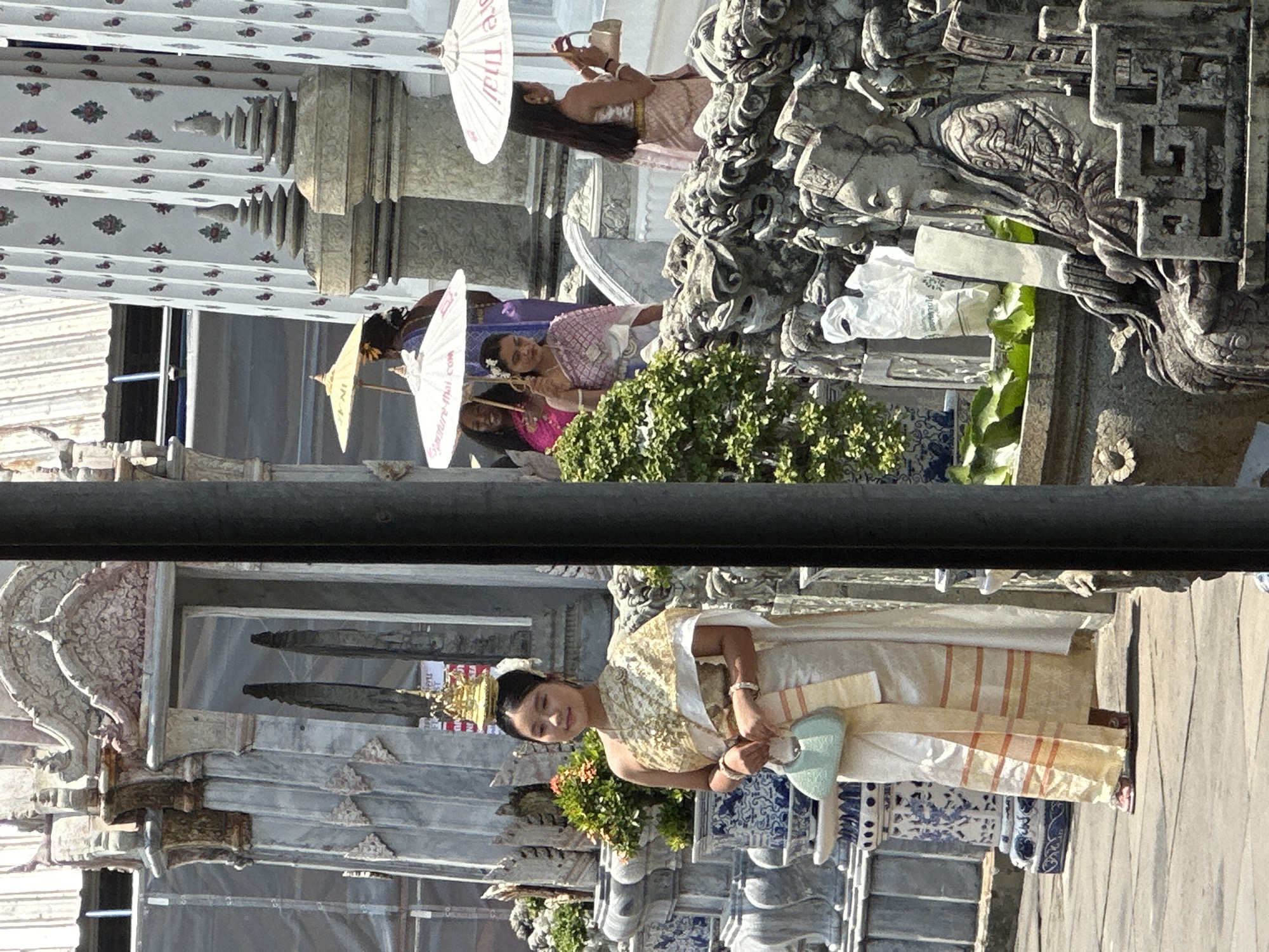 The image depicts a scene at Wat Arun Ratchawararam Ratchawaramahawihan (Temple of Dawn) in Bangkok, Thailand. A woman in traditional Thai dress stands in the foreground. Behind her is the temple architecture, featuring intricate ornamentation and tiled surfaces. Three women in traditional dress, each holding a white paper parasol, are captured in the background near a stone sculpture.