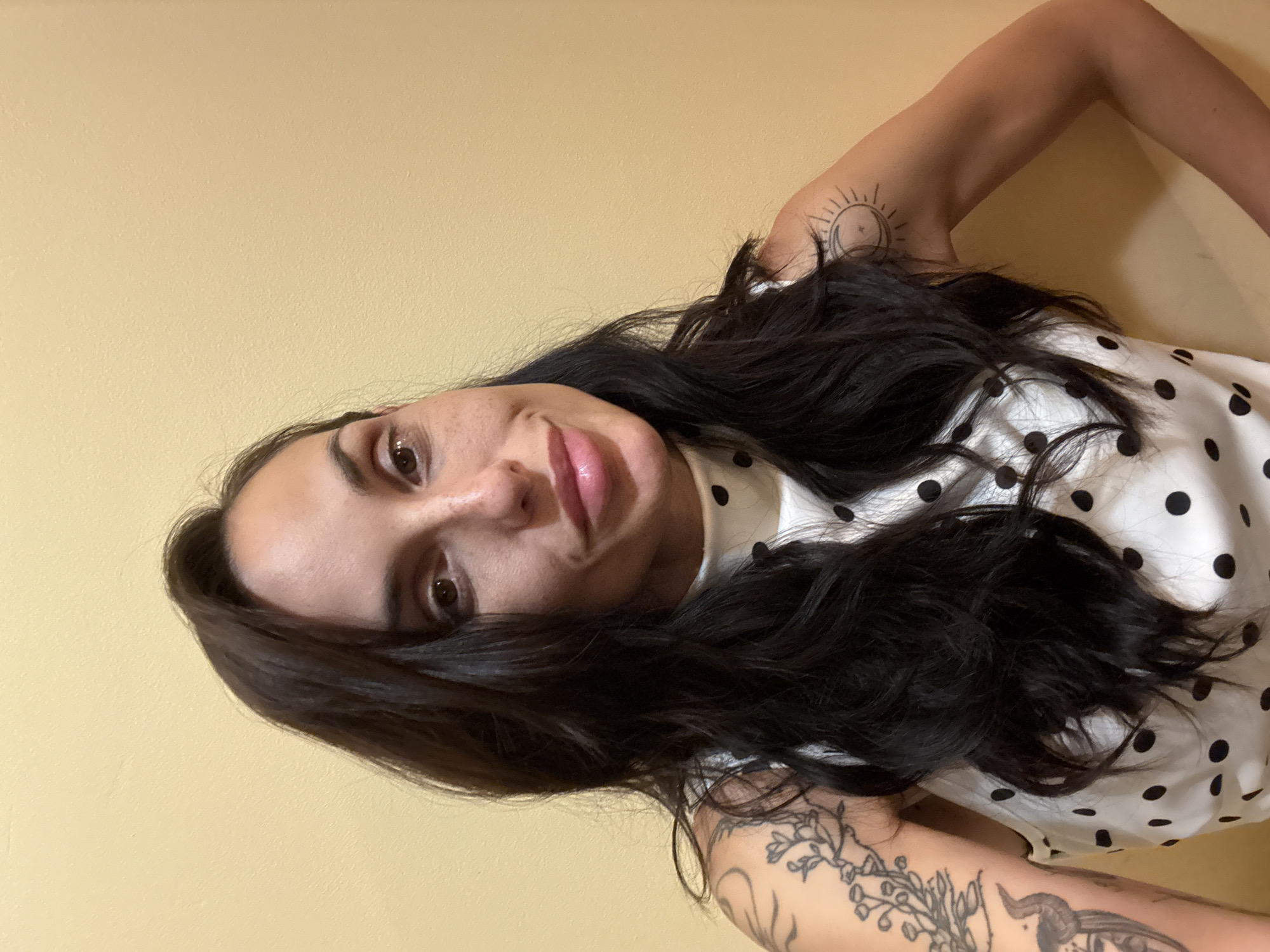 A woman is featured in a portrait photograph, oriented 90 degrees clockwise. She has long, dark, wavy hair that cascades around her. Her gaze is directed towards the camera, accompanied by a subtle, relaxed smile. Her eyes are dark brown, complemented by visible eye makeup, and her lips have a glossy, pink hue.
She is dressed in a white top patterned with black polka dots. Both of her arms display tattoos. On her left upper arm (viewer's right), there is a distinct tattoo featuring a sun and moon design. On her right arm (viewer's left), an intricate floral or branch-like tattoo is visible, extending downwards, with another partial tattoo, possibly an animal head (such as a goat or bull), visible further along the arm.
The background consists of a plain, solid light yellow wall, suggesting an indoor setting. The lighting is soft and even. No text is visible in the image. The overall impression is one of calm and gentle contemplation. The provided context for the image is Pleven, Bulgaria.